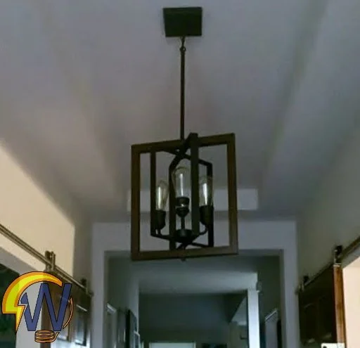 Modern pendant light with a rectangular wooden frame and three candle-style bulbs hanging from the ceiling in a hallway.