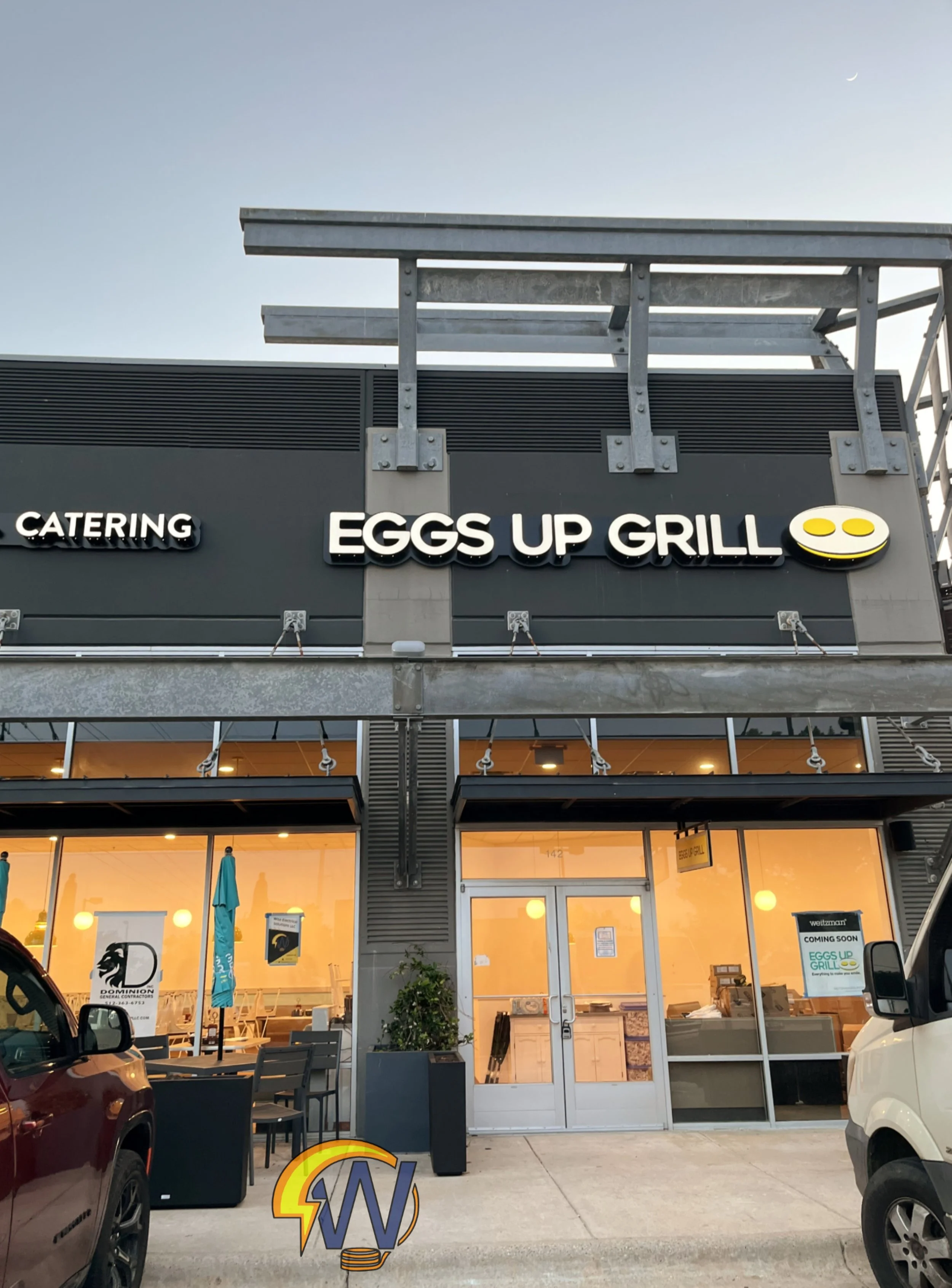 A commercial storefront named 'Eggs Up Grill' with outdoor seating and a partially visible sign for a different business called 'D' and another sign indicating 'Coming Soon.' The facade features large glass windows showing the interior, with warm lig