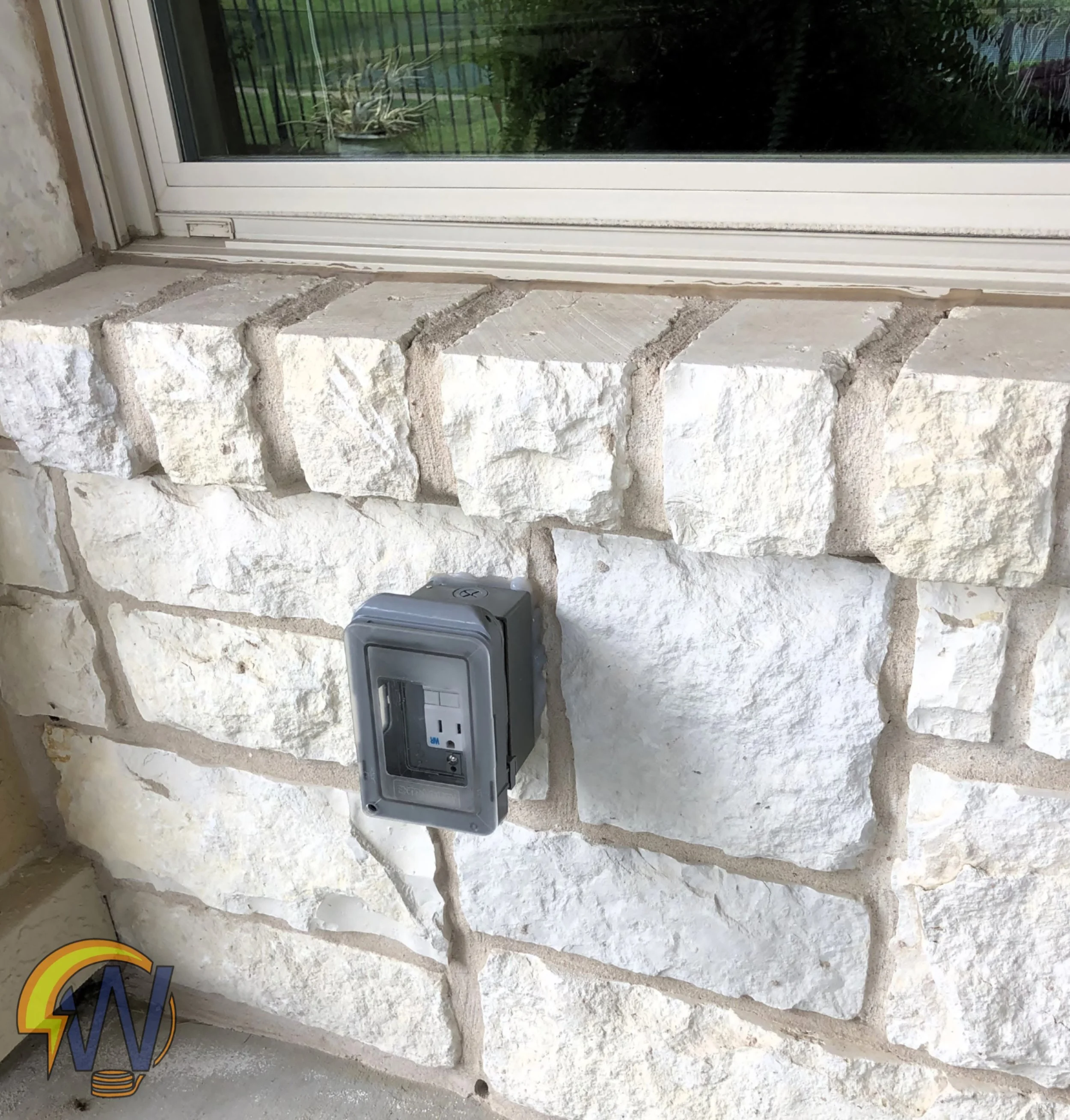 A stone wall with an electrical outlet box attached, and a window with a view of green trees and plants outside.