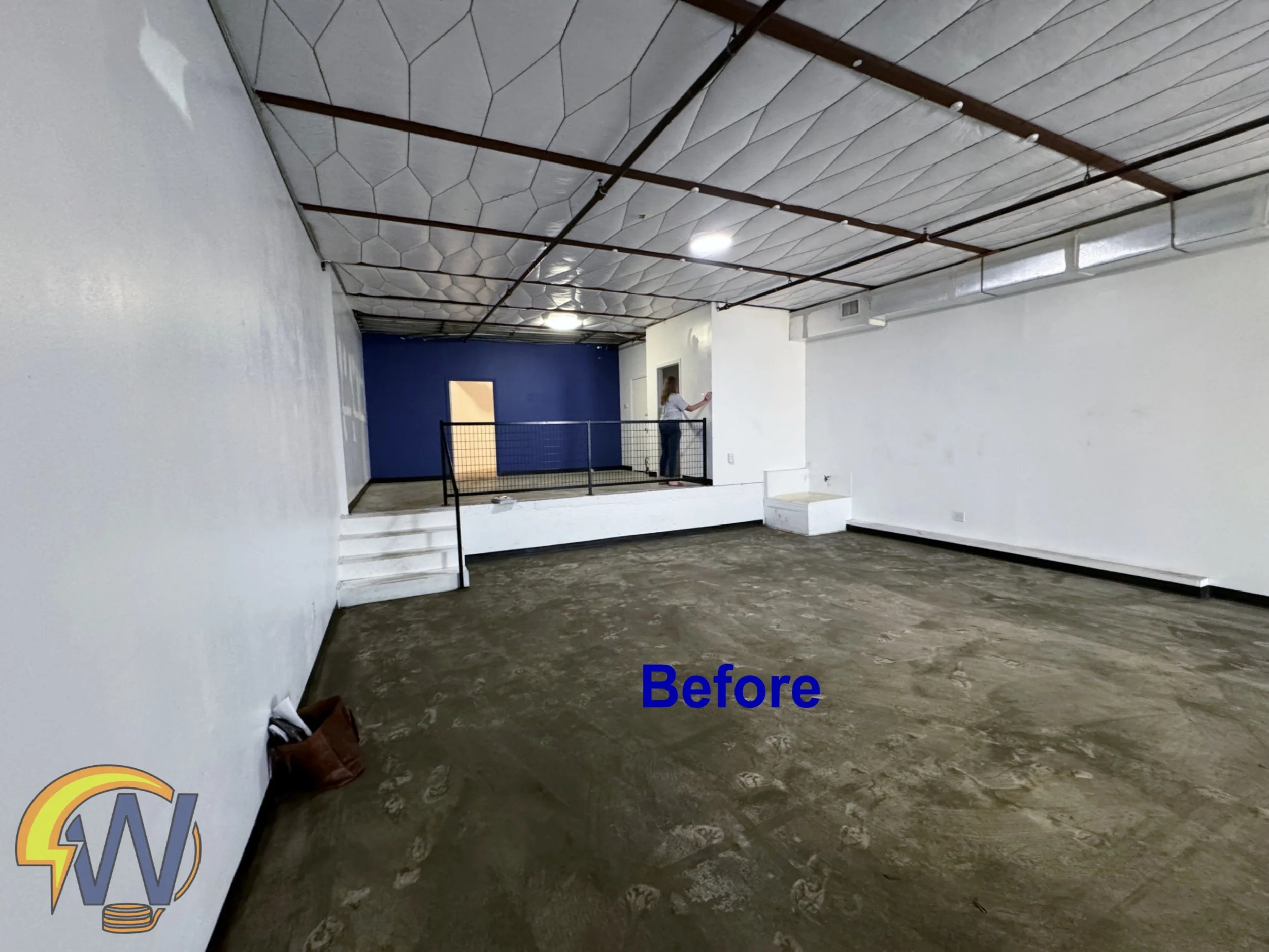 An empty room with white walls, a blue accent wall at the back, and a brown concrete floor. There are steps leading up to a small platform with a woman standing on it, writing on the wall. The ceiling has a geometric pattern with white panels and exp