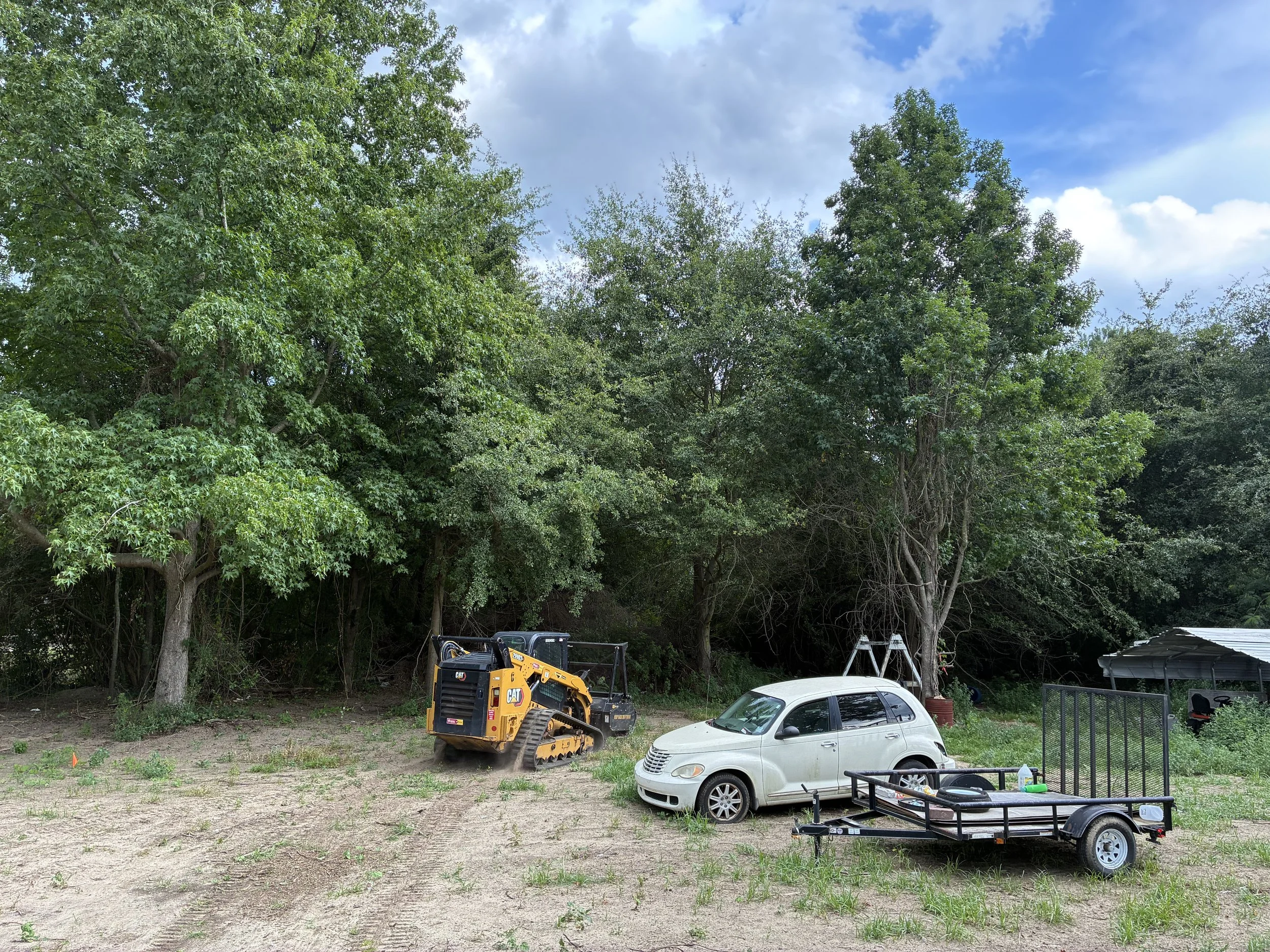 A backyard with trees, a small white car, a yellow construction vehicle, a trailer, and patio furniture.