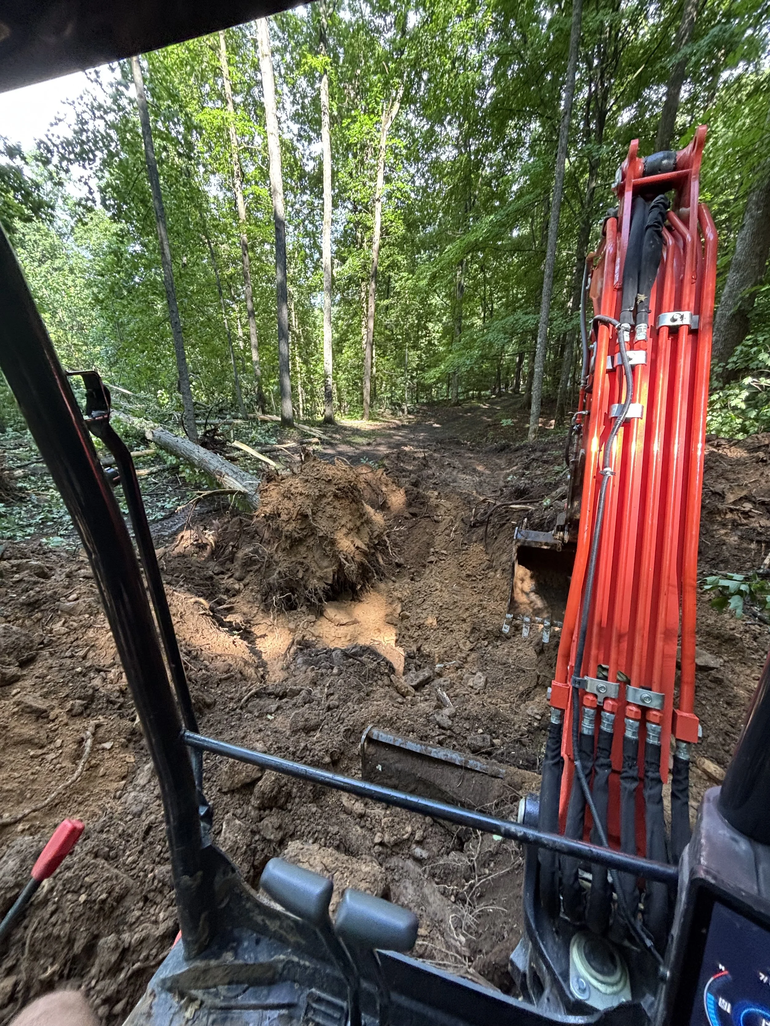 View from inside a construction vehicle cab looking out into a forested area with trees and dirt. The vehicle's control panel and red hydraulic arm are visible.