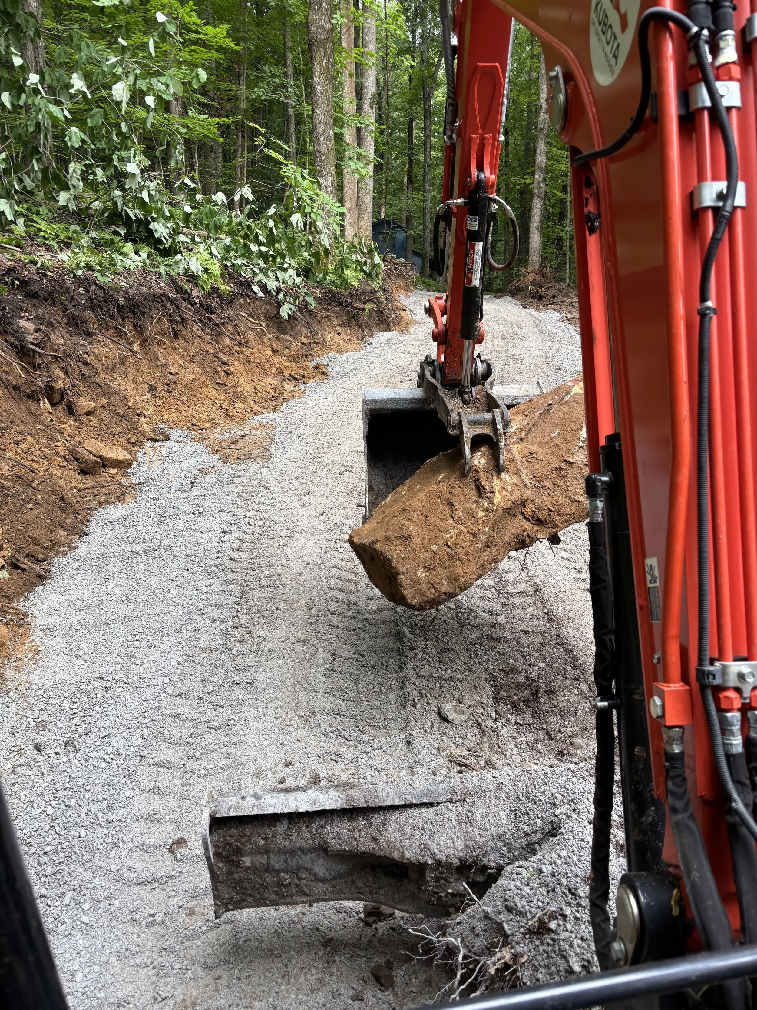 A construction excavator is removing large rocks and dirt from a forest trail, creating a tunnel-like hole through the ground.