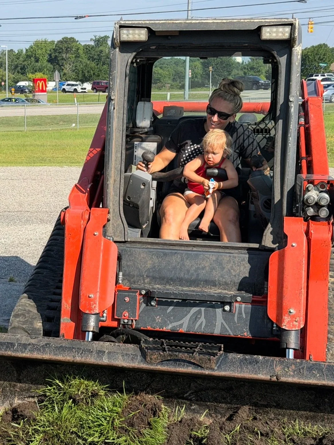 A woman and a young girl sitting inside a red construction vehicle, possibly a skid-steer loader, with the woman smiling and the girl looking slightly serious, wearing sunglasses and casual summer clothes.
