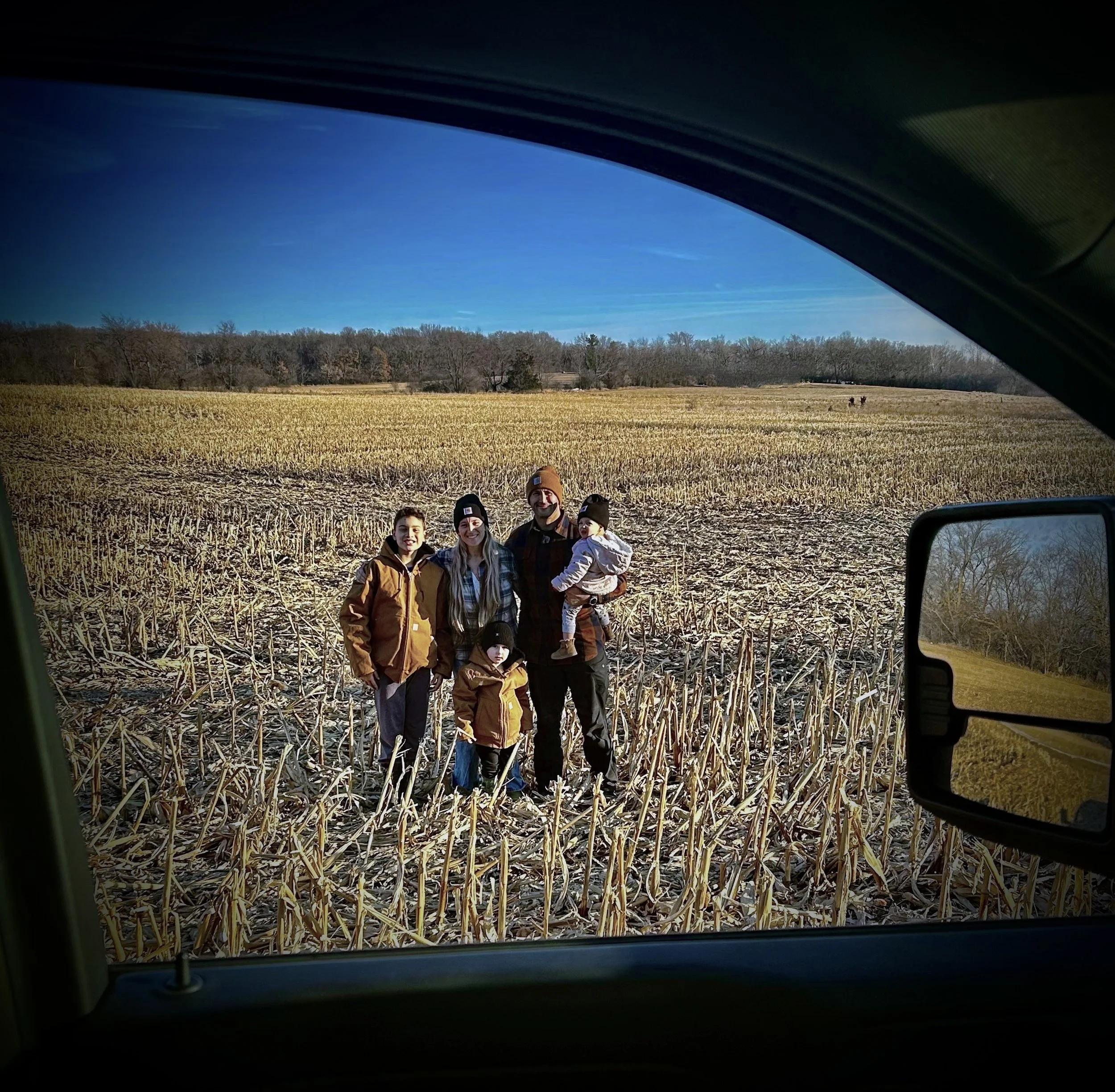 Photo taken from inside a vehicle showing a group of five people standing in a harvested field with trees and a clear blue sky in the background.