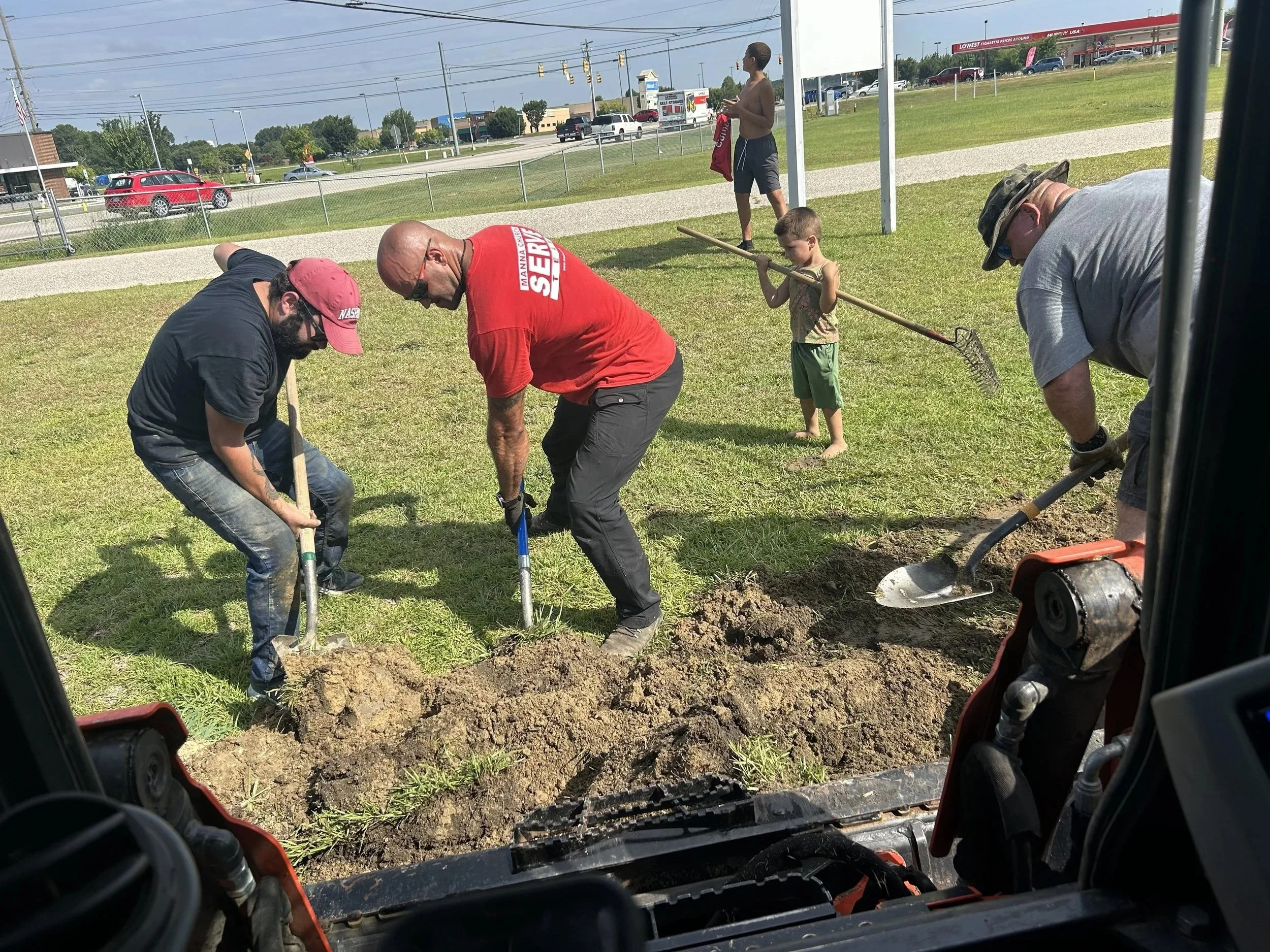 Group of men and a young boy working together digging a hole in a grassy area for planting or construction, viewed from inside a construction vehicle.
