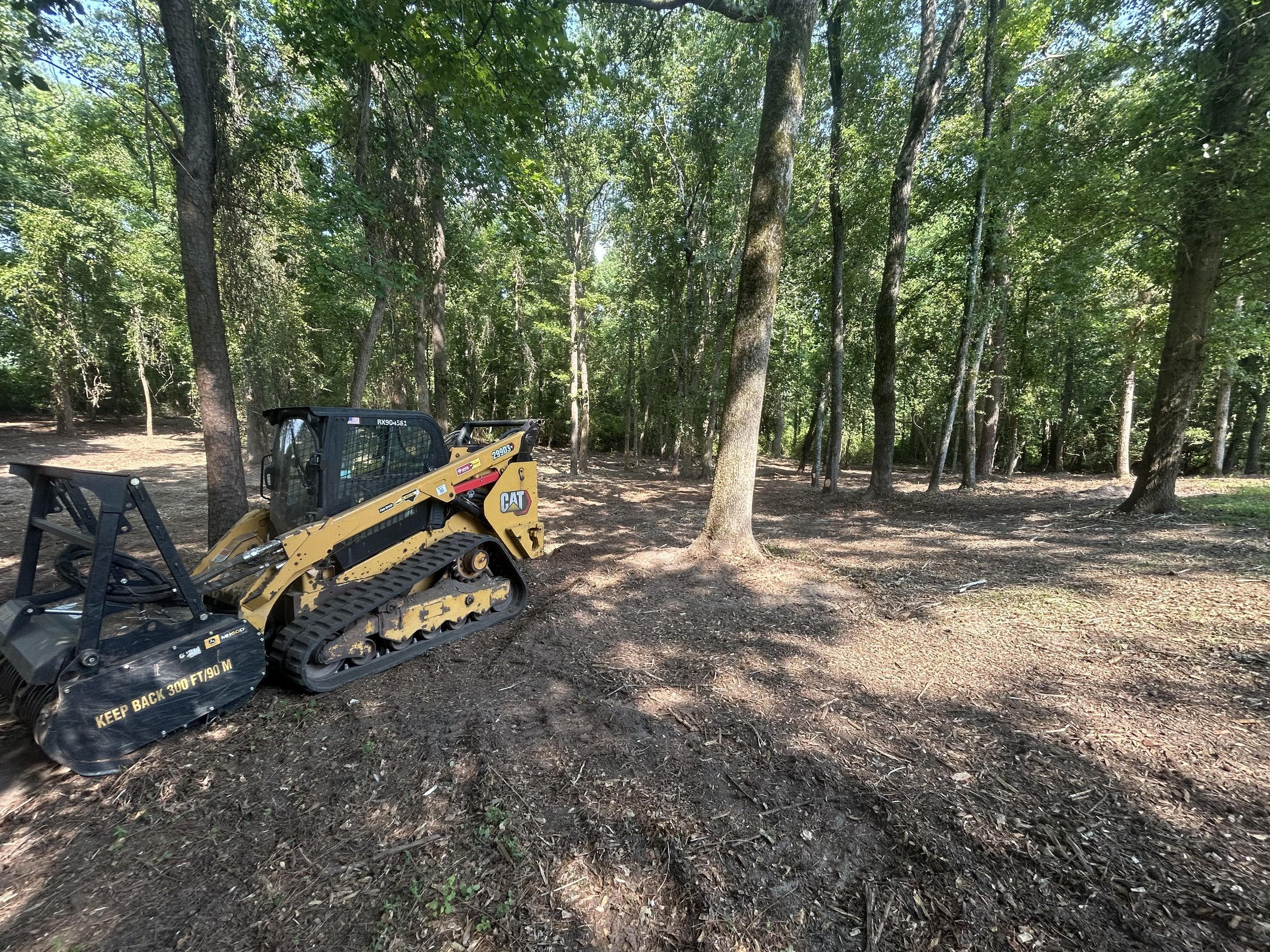 A wooded area with trees and dirt ground, and a small construction vehicle with tracks and a cutting blade.