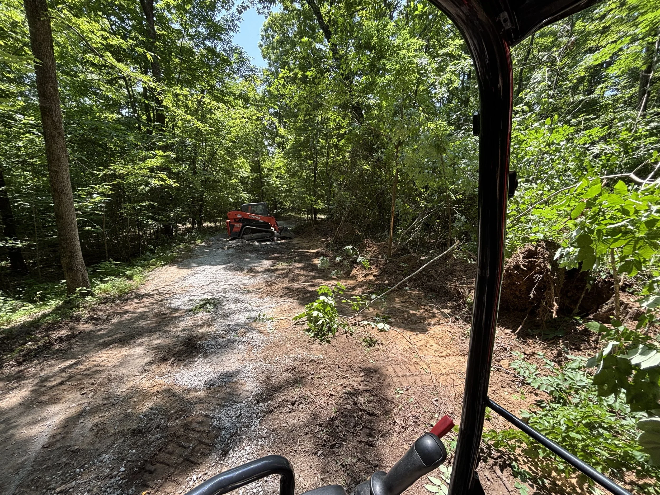 View from the inside of a small vehicle or machinery cabin looking out onto a narrow dirt trail surrounded by dense green forest. A small red and black tracked vehicle is visible on the trail.