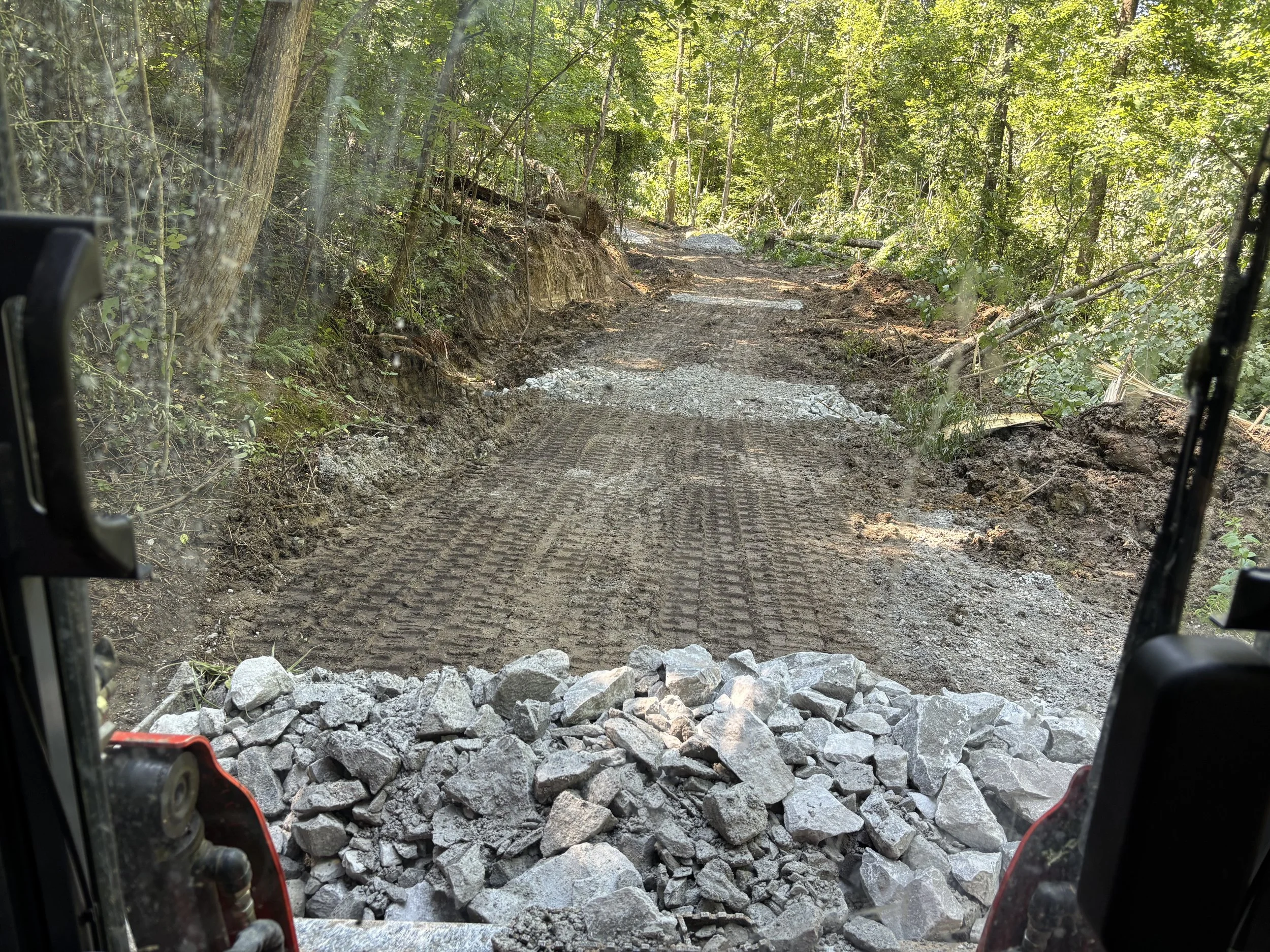 View from inside a construction vehicle looking out onto a dirt and gravel road in a forest, with rocks and dirt in the foreground.