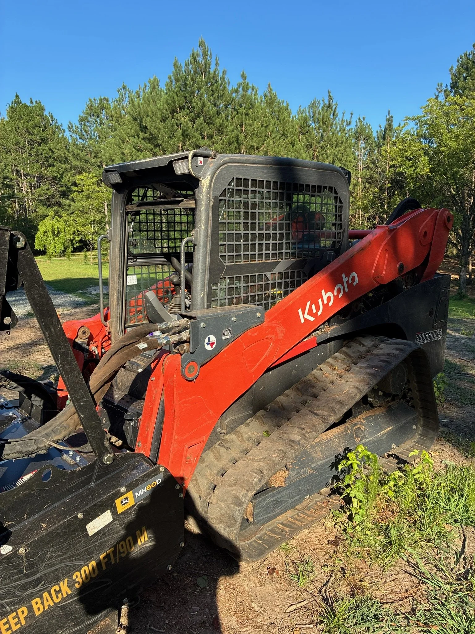 Close-up of a Kubota tractor with tracks, parked on a grassy area with trees and a clear blue sky in the background.