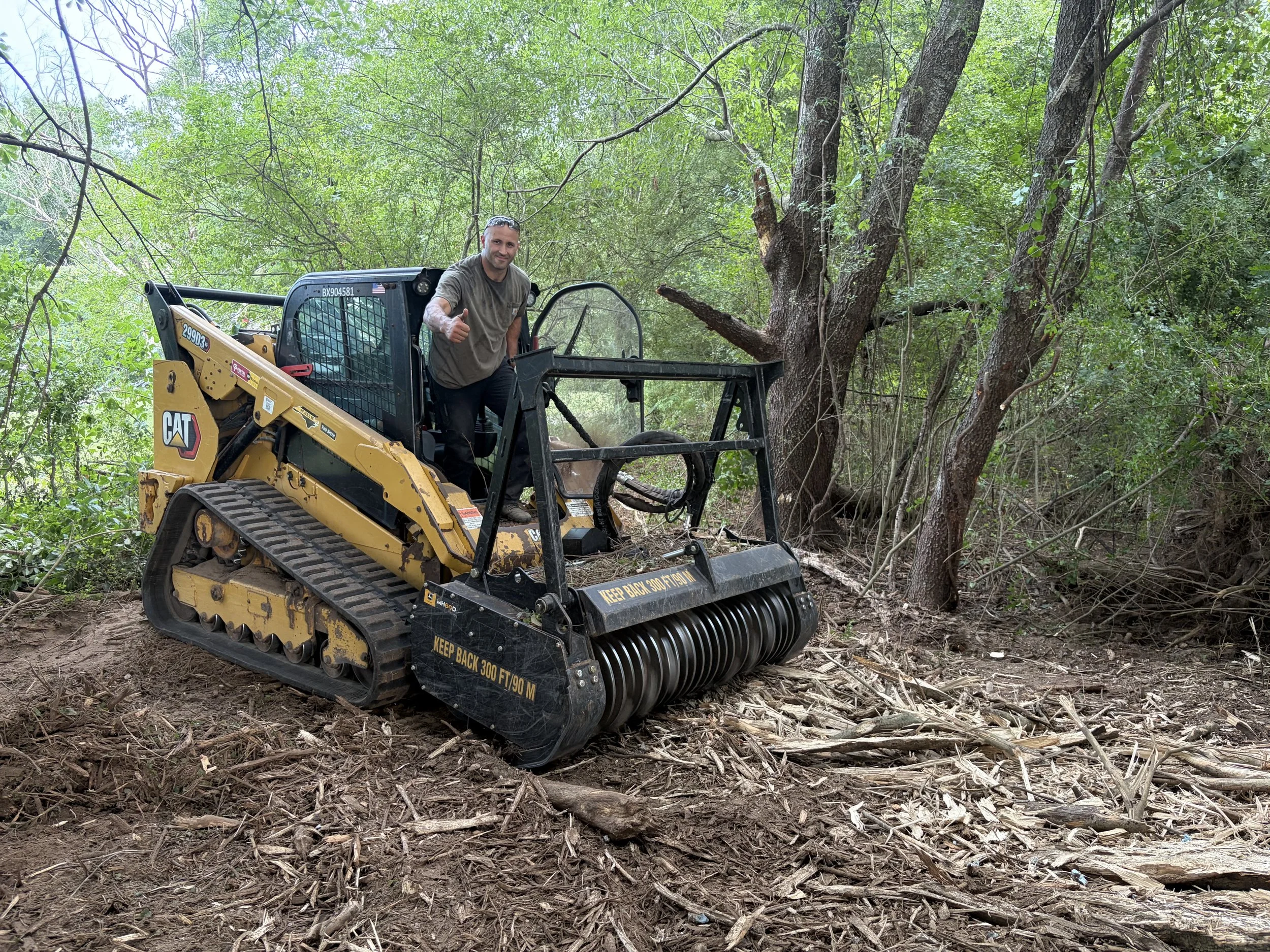 A man operating a small tracked bulldozer or construction vehicle in a wooded area with green leaves and trees, giving a thumbs-up gesture.