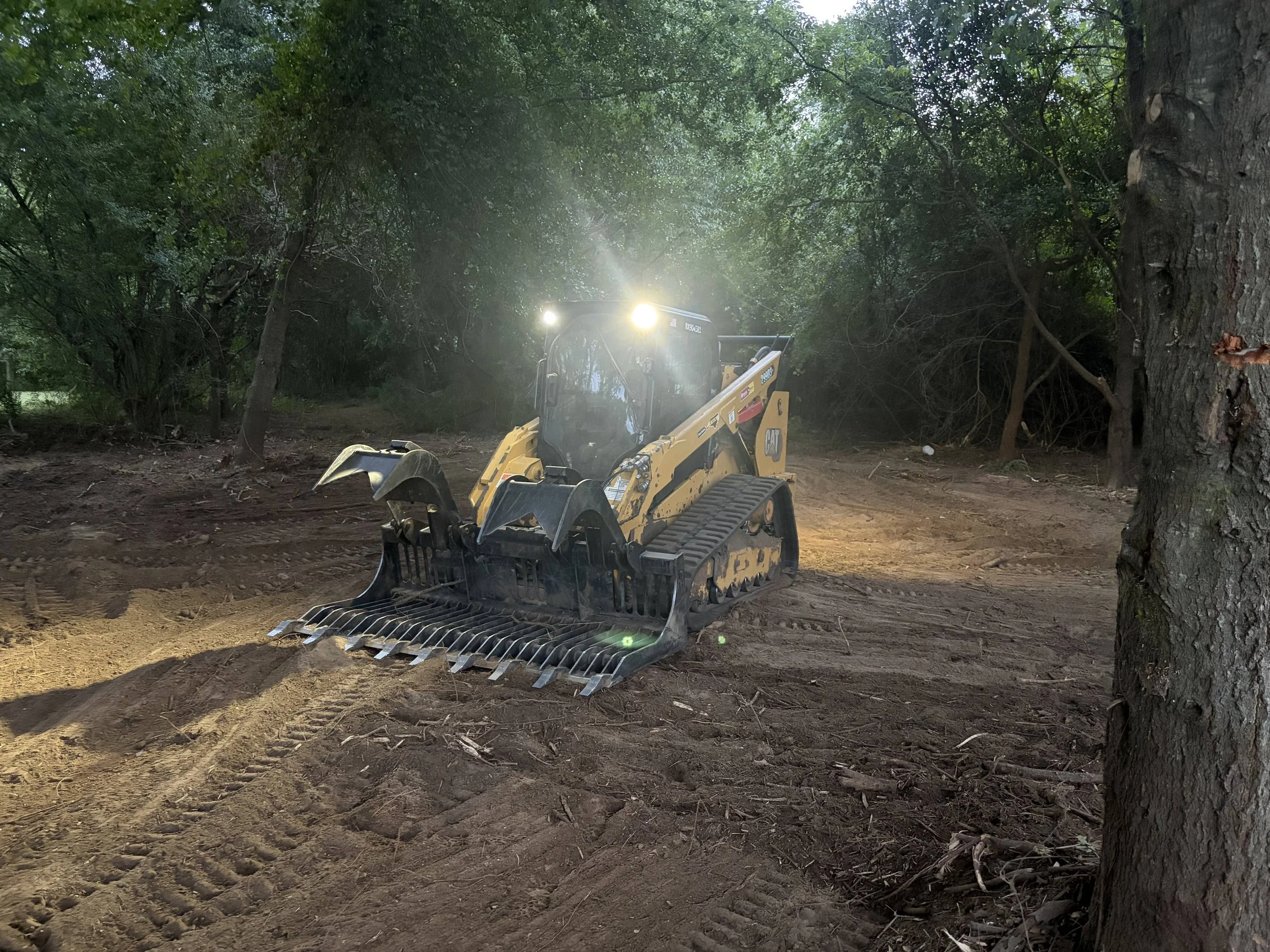 A construction vehicle working on clearing trees and dirt in a wooded area, with sunlight filtering through the leaves.