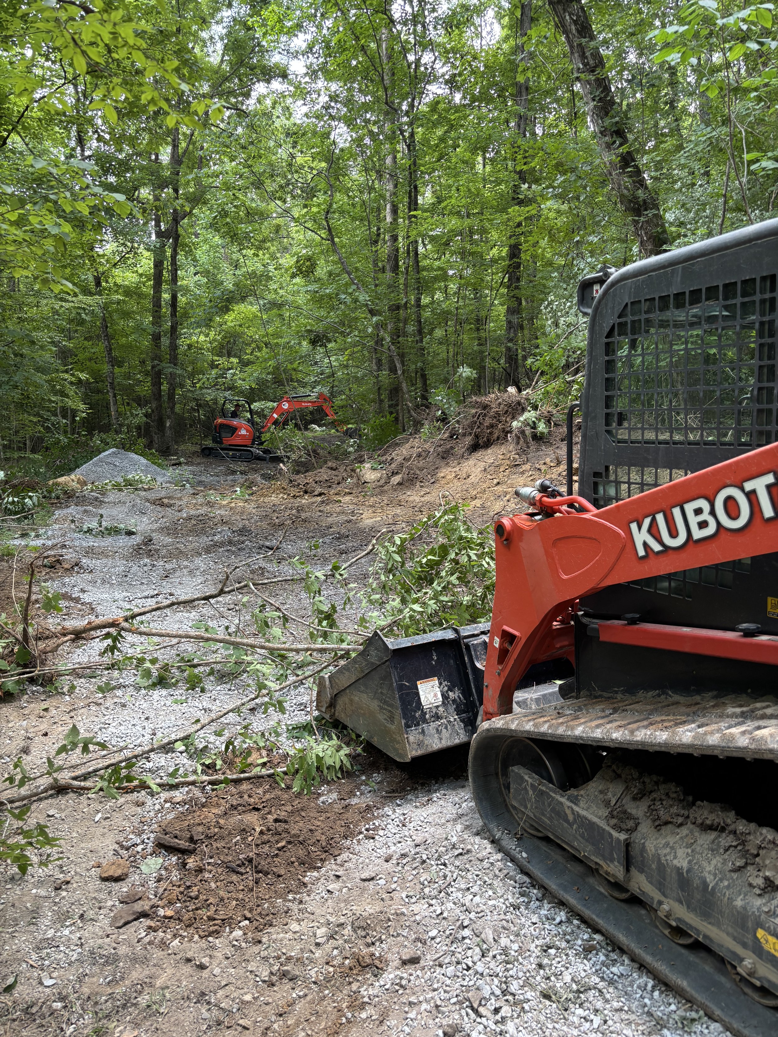 A small orange excavator working on a dirt and gravel trail in a dense green forest.