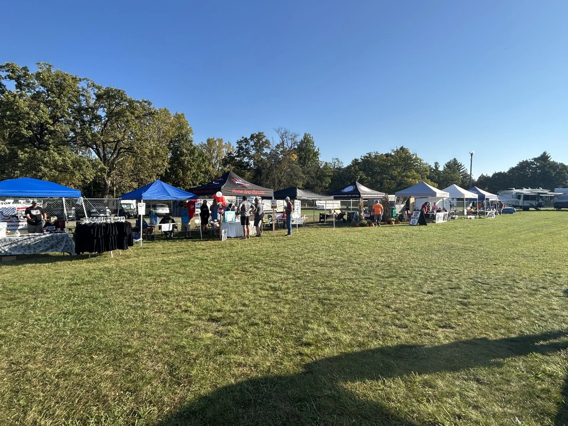 Outdoor fair with various tents and booths set up on a grassy field, with people browsing and socializing under a clear blue sky.