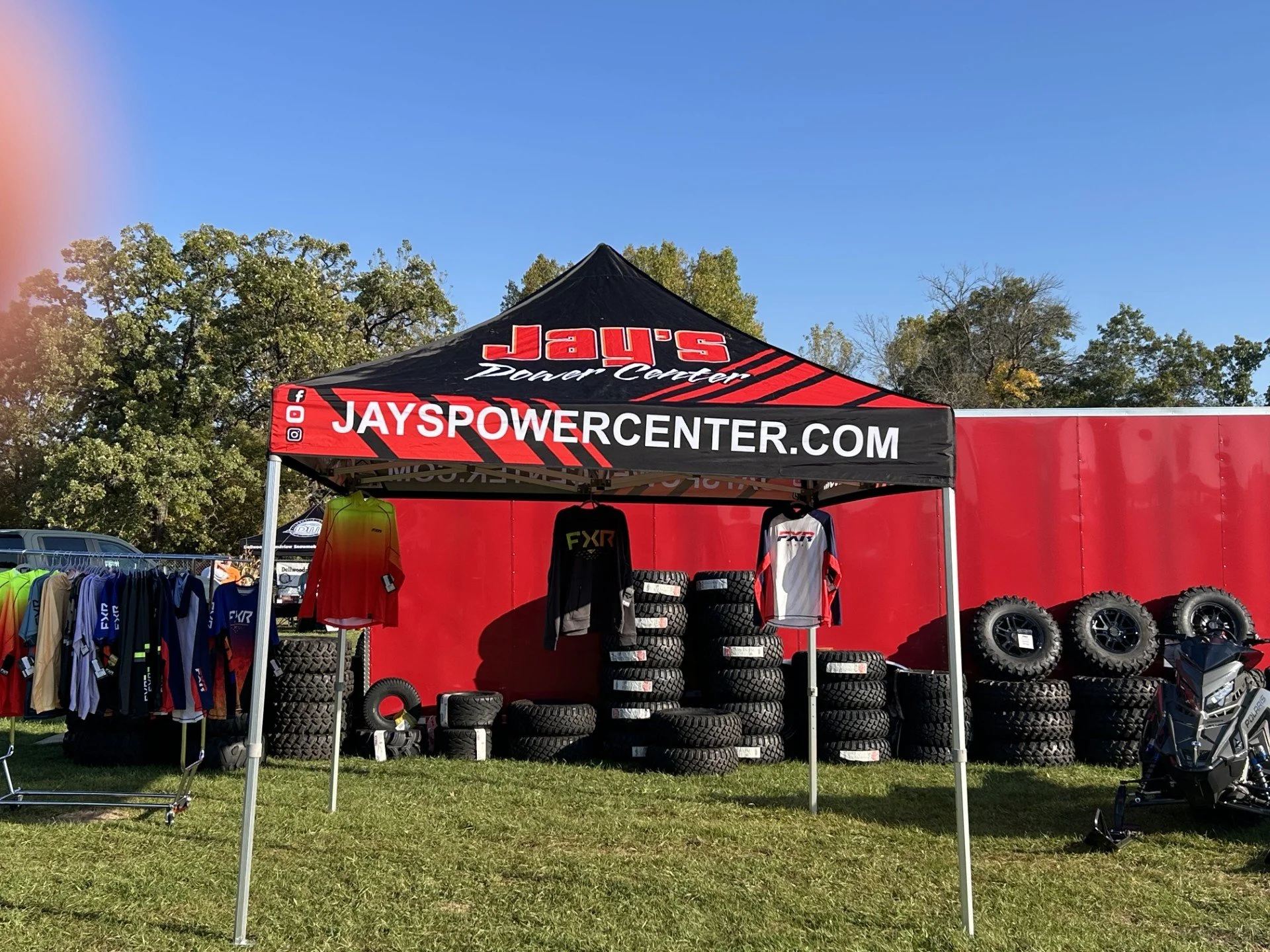Outdoor tent with promotional signage for JAYS Power Center, displaying tires, apparel, and racing equipment in a grassy area with trees and a clear blue sky.