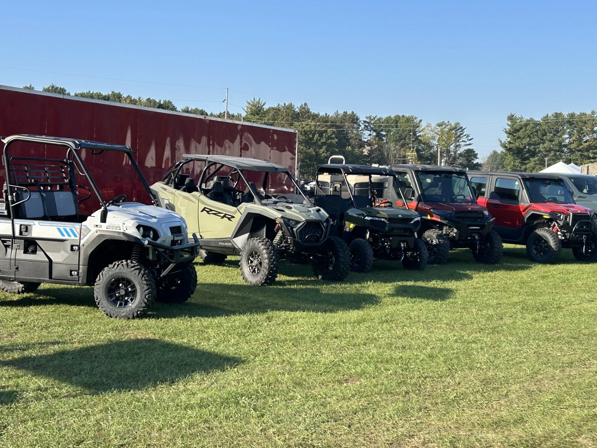 Line of off-road utility vehicles parked on grassy field with red trailer and trees in background.