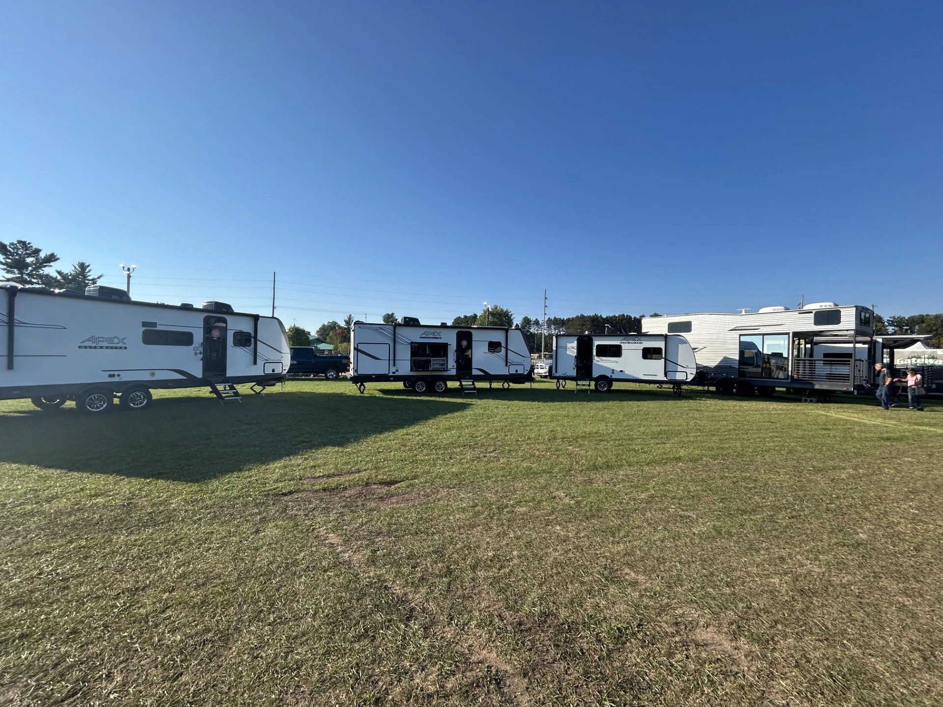 Multiple recreational vehicles and trailers parked on a grassy field under a clear blue sky, with trees and power lines in the background.