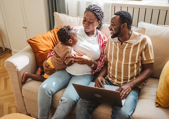 A family of three sitting on a beige couch in a living room, with a laptop on the man's lap. The woman is holding a young child on her lap, and they are all smiling at each other.