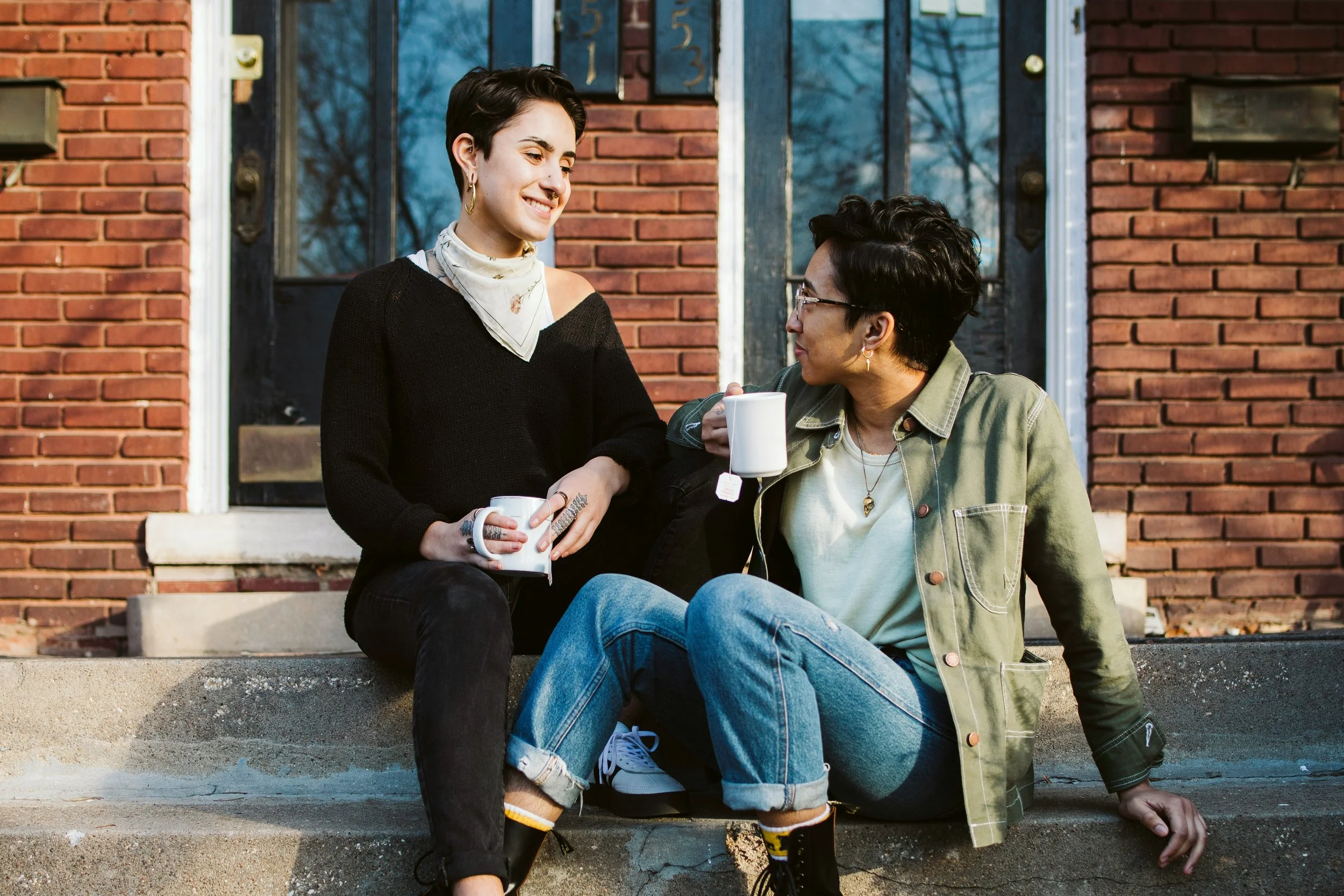 Two women sitting on the steps of a house, smiling, holding coffee mugs, and engaging in conversation outdoors on a sunny day.