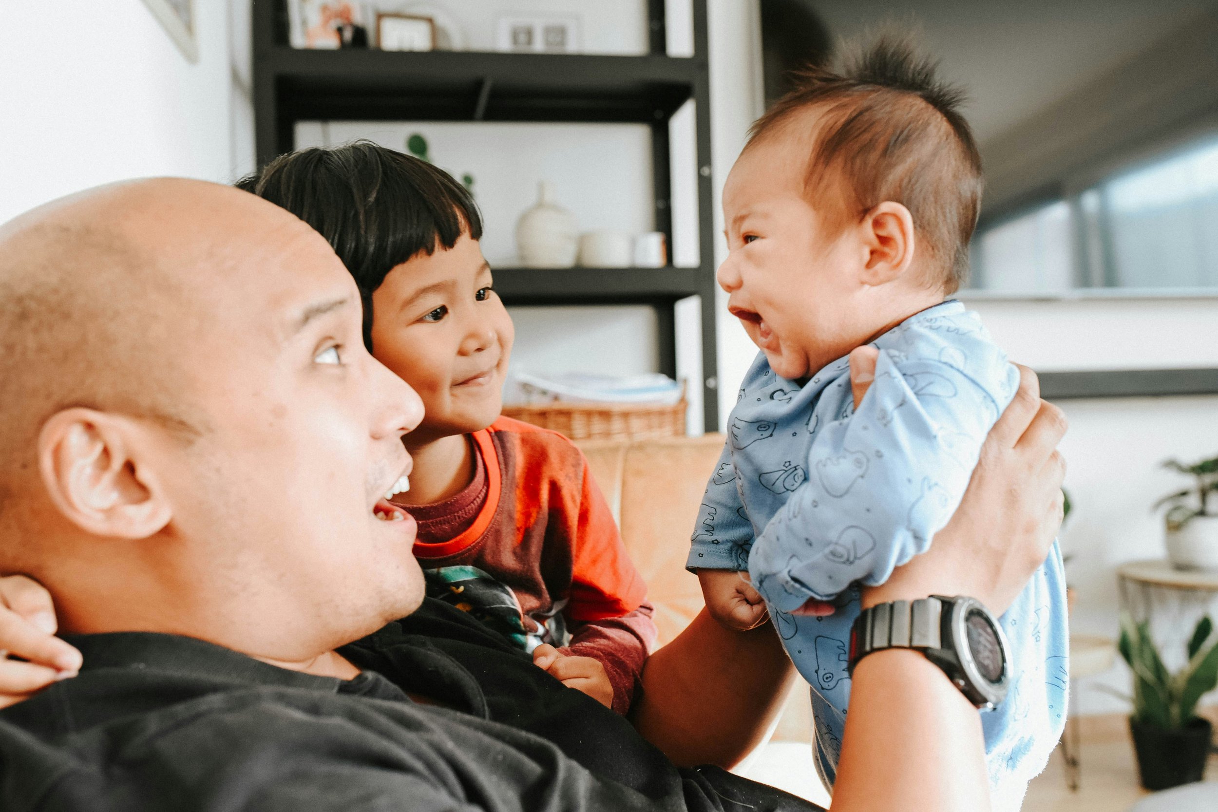 An adult holding a baby with one hand and a young girl with the other, all facing each other indoors, smiling and engaging at the camera.