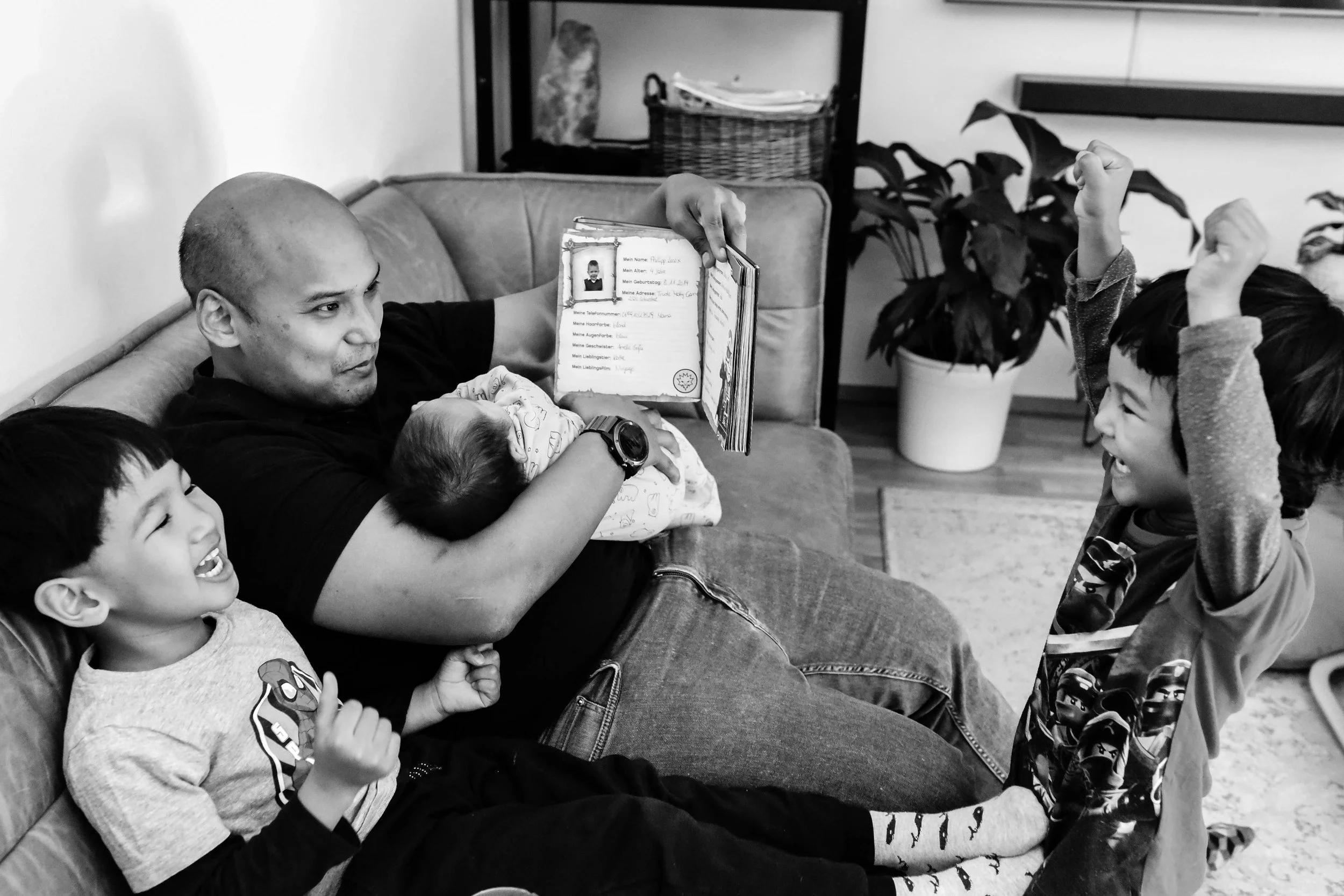 A man holding a baby and sitting on a couch with children around, some raising their hands, in a room with potted plants and shelves.