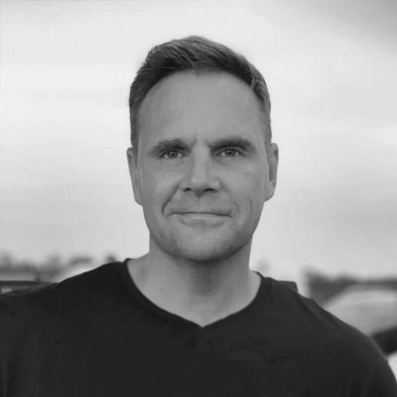 A black-and-white photo of a man with short hair, wearing a black shirt, outdoors with a cloudy sky in the background.