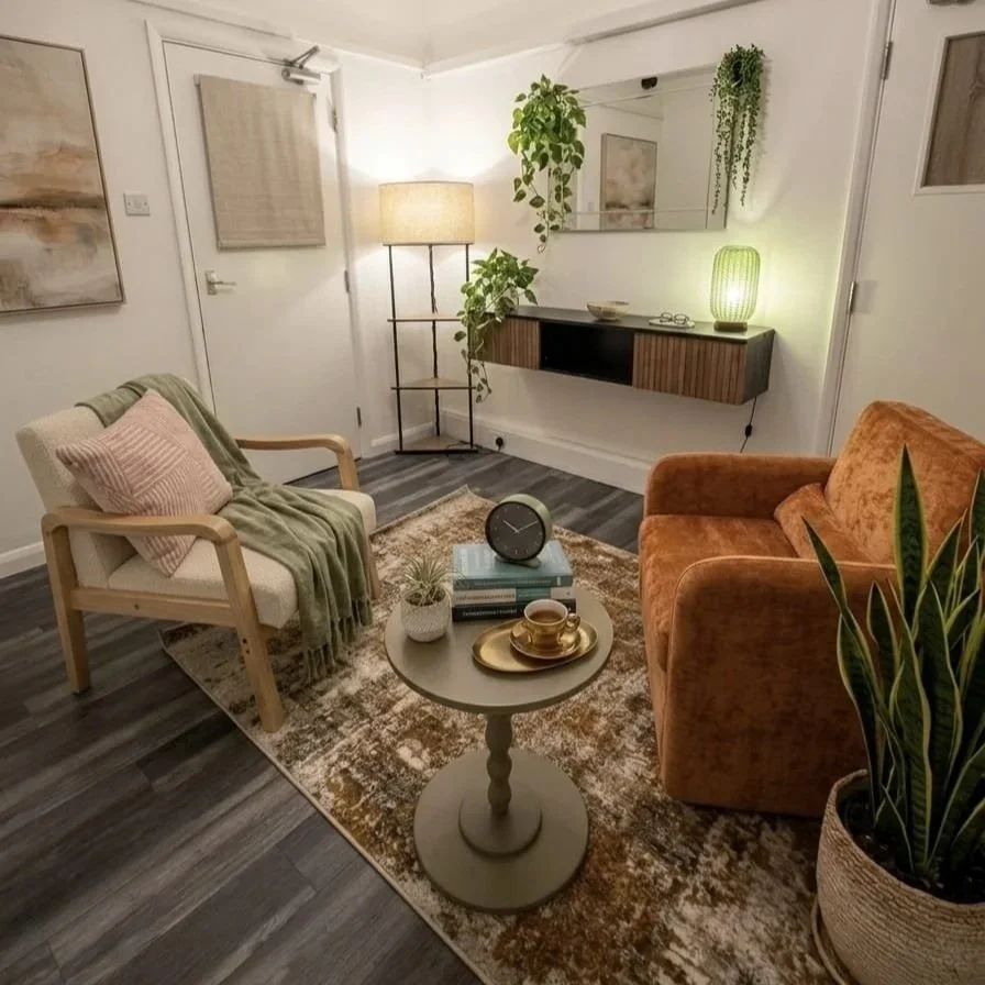 Cozy living room with a white armchair draped with a green blanket and pink cushion, an orange sofa, a round coffee table with books, a clock, and cups, a striped rug, a tall floor lamp, wall hanging, wall mirror, floating shelf with decorations, and potted houseplants.