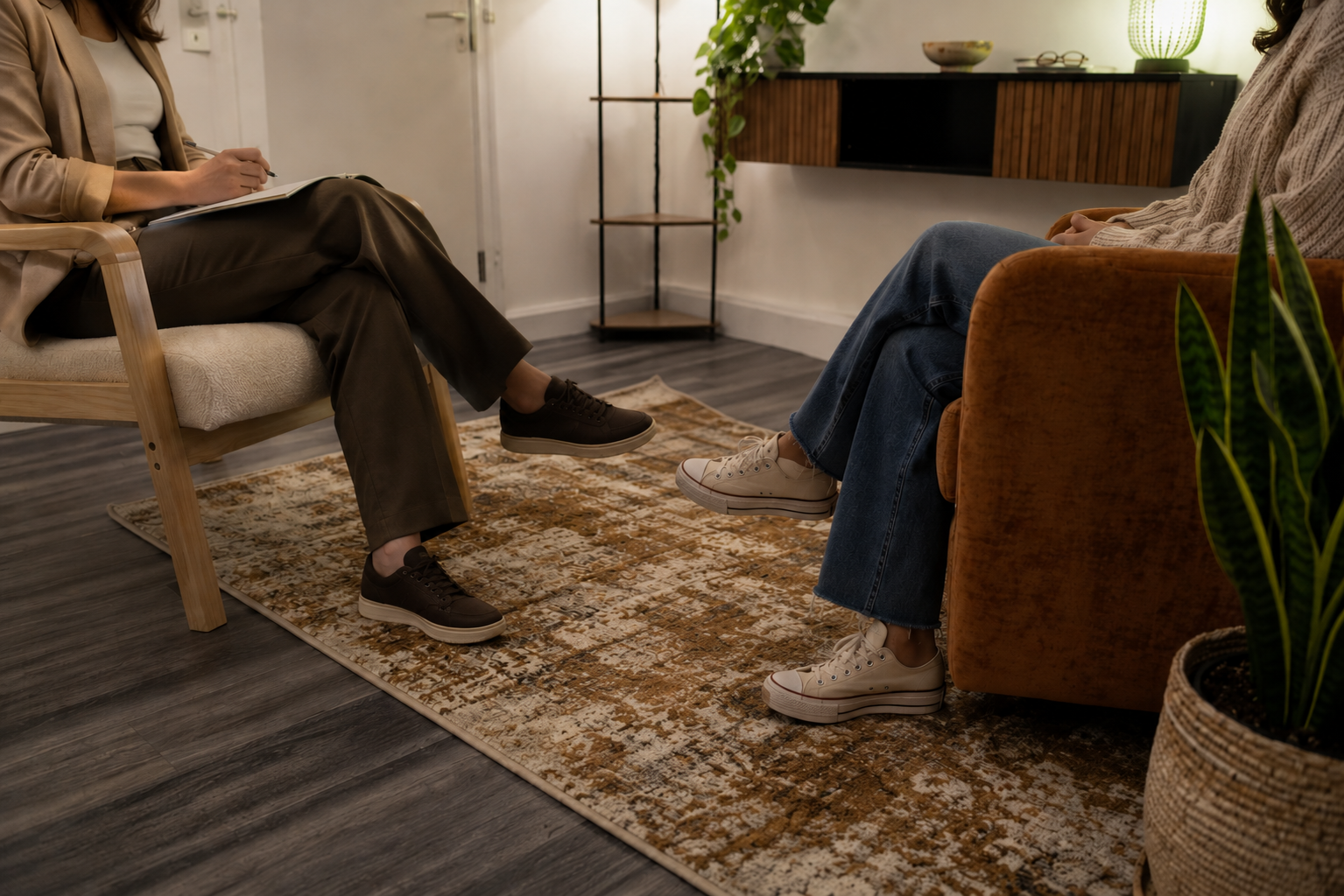 A therapy session with two women sitting across from each other in a cozy, well-lit room. Both are wearing casual shoes, and the room has a patterned rug, plants, and minimal decor.