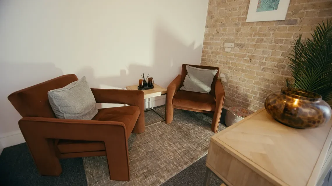 Cozy living room corner with two brown armchairs, a small wooden side table, a textured area rug, a brick accent wall, and decorative items including a large plant in a brown vase and framed artwork on the wall.