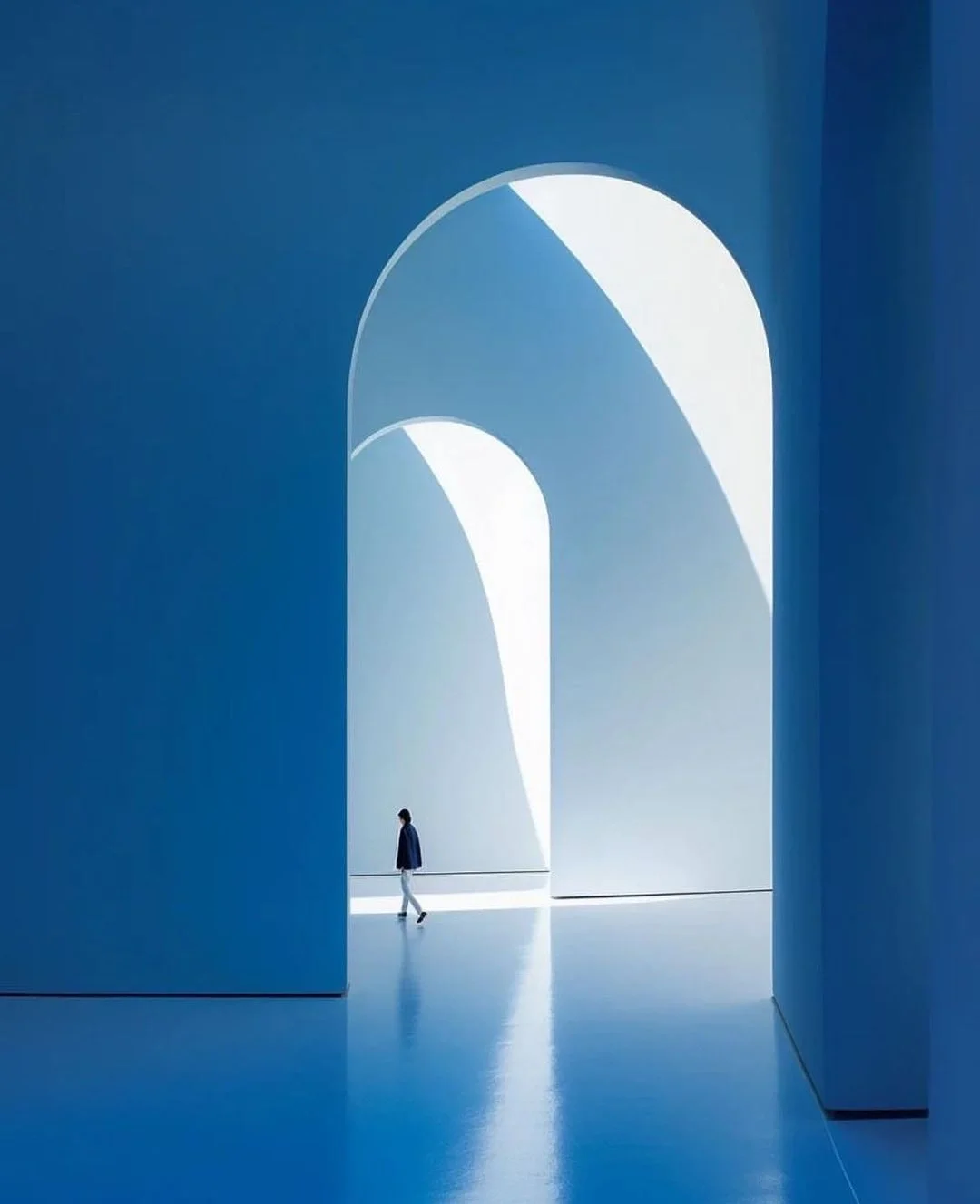 A person walking through a modern, minimalist architectural space with large, arched openings and blue walls, illuminated by natural light.