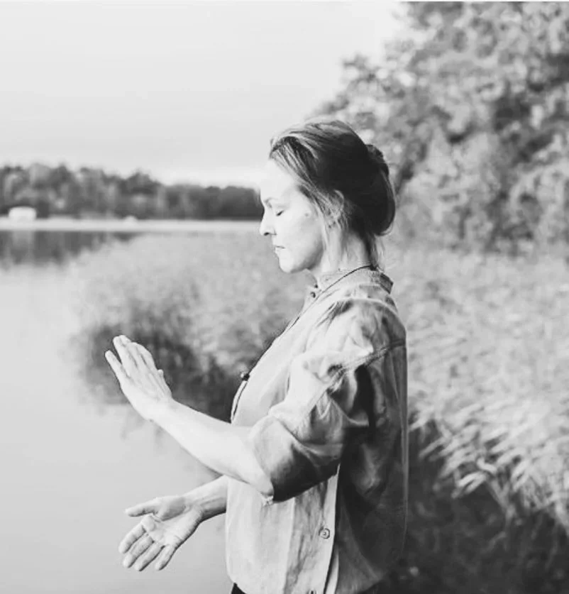 A woman standing near a lake with her hands pressed together in a prayer or meditative posture, facing slightly downward with a serene expression.
