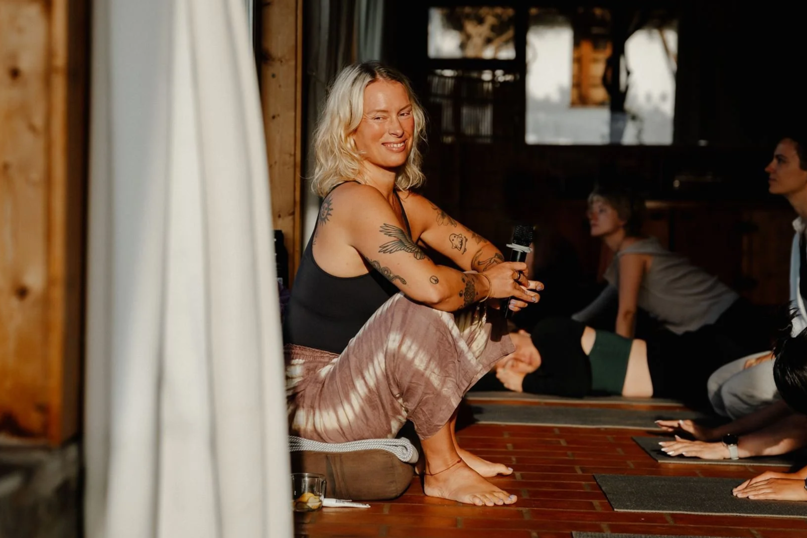 A woman with blonde hair and tattoos on her arms sitting on the floor holding a microphone during a yoga or meditation session in a rustic wooden room, with other participants in the background.