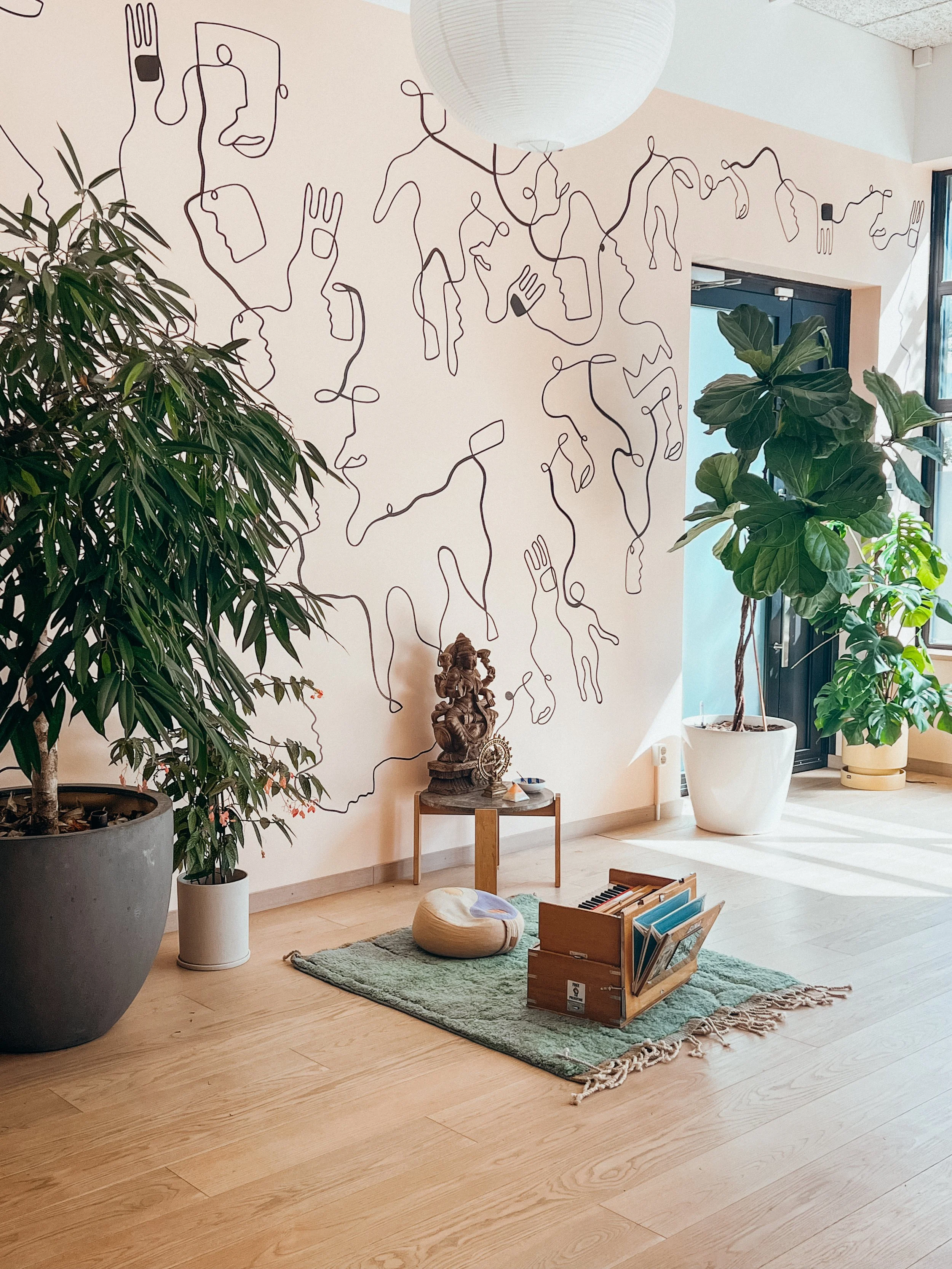 A cozy interior space with large potted plants, a green rug, a small wooden table with a decorative sculpture, and a wall featuring minimalist line art of faces and hands. There is a round paper lantern hanging from the ceiling and sunlight coming through a window.