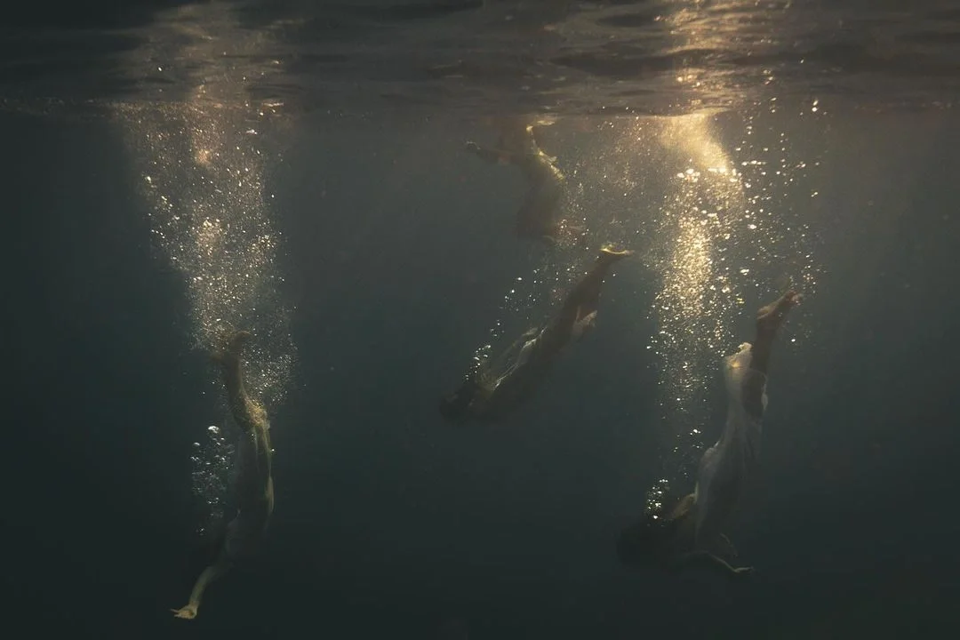 Three people diving into a body of water, creating bubbles and ripples, with sunlight reflecting off the water's surface.