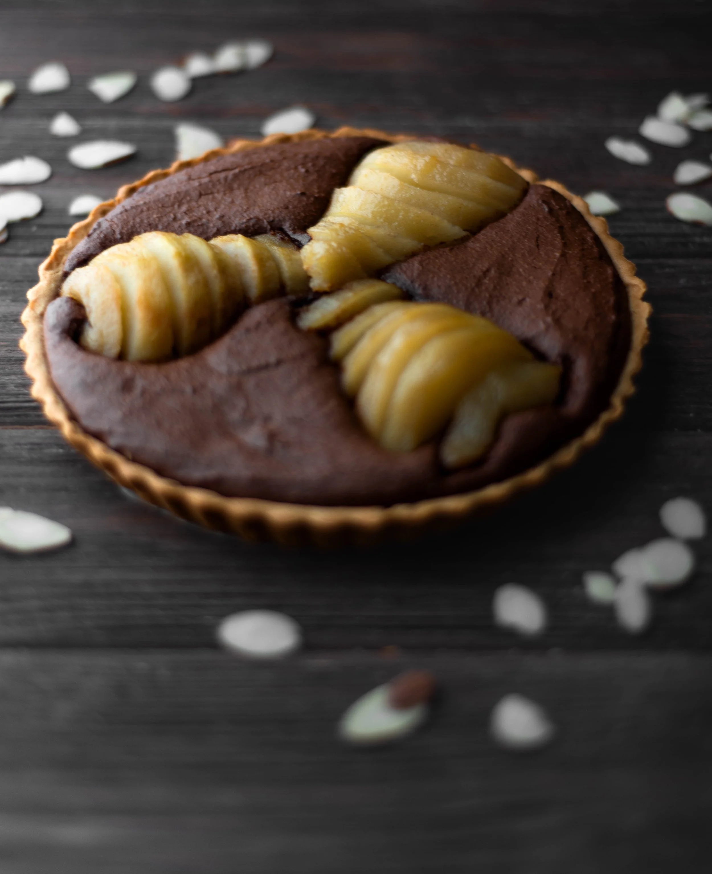 A tart with pastry crust, filled with dark chocolate and topped with sliced bananas, on a dark wooden surface with scattered white and brown seeds or nuts.