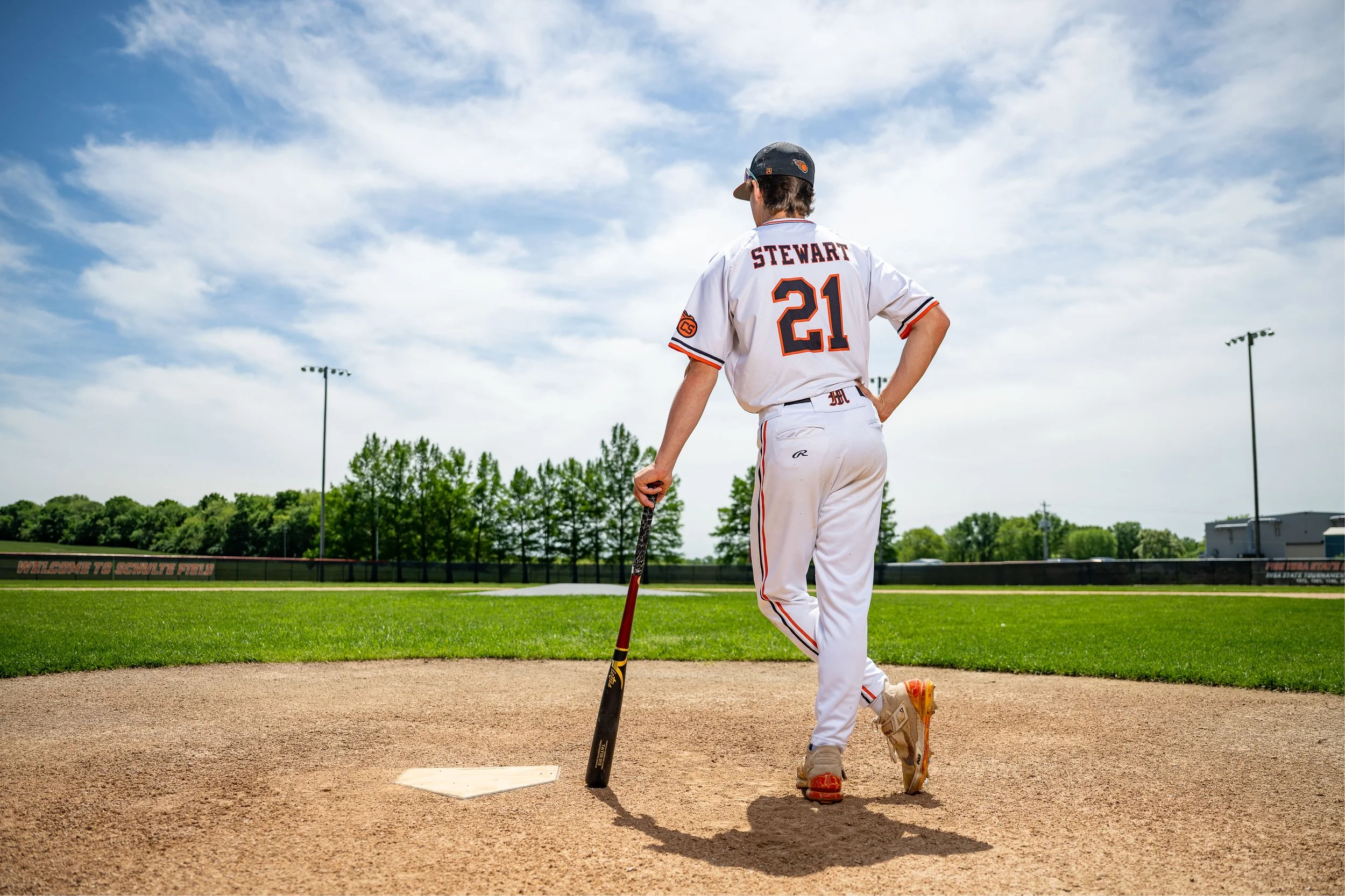 A baseball player standing on a baseball field, viewed from behind. He is wearing a white uniform with black and orange accents, a cap, and holding a bat. The game is outdoors on a bright day with a partly cloudy sky, and resoding trees and stadium lights in the background.