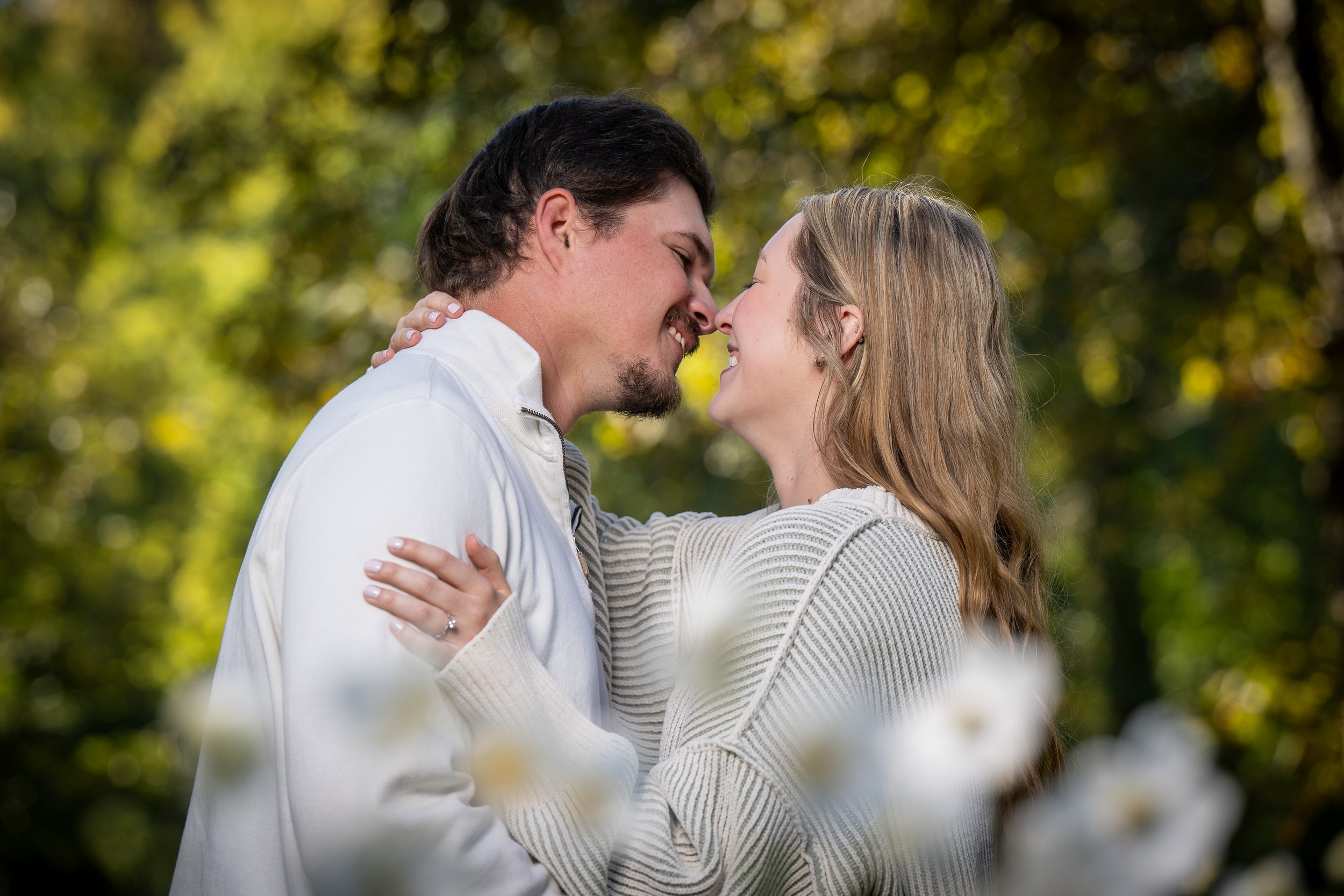 A young man and woman face each other with noses touching, smiling, outdoors with blurred trees and sunlight in the background.