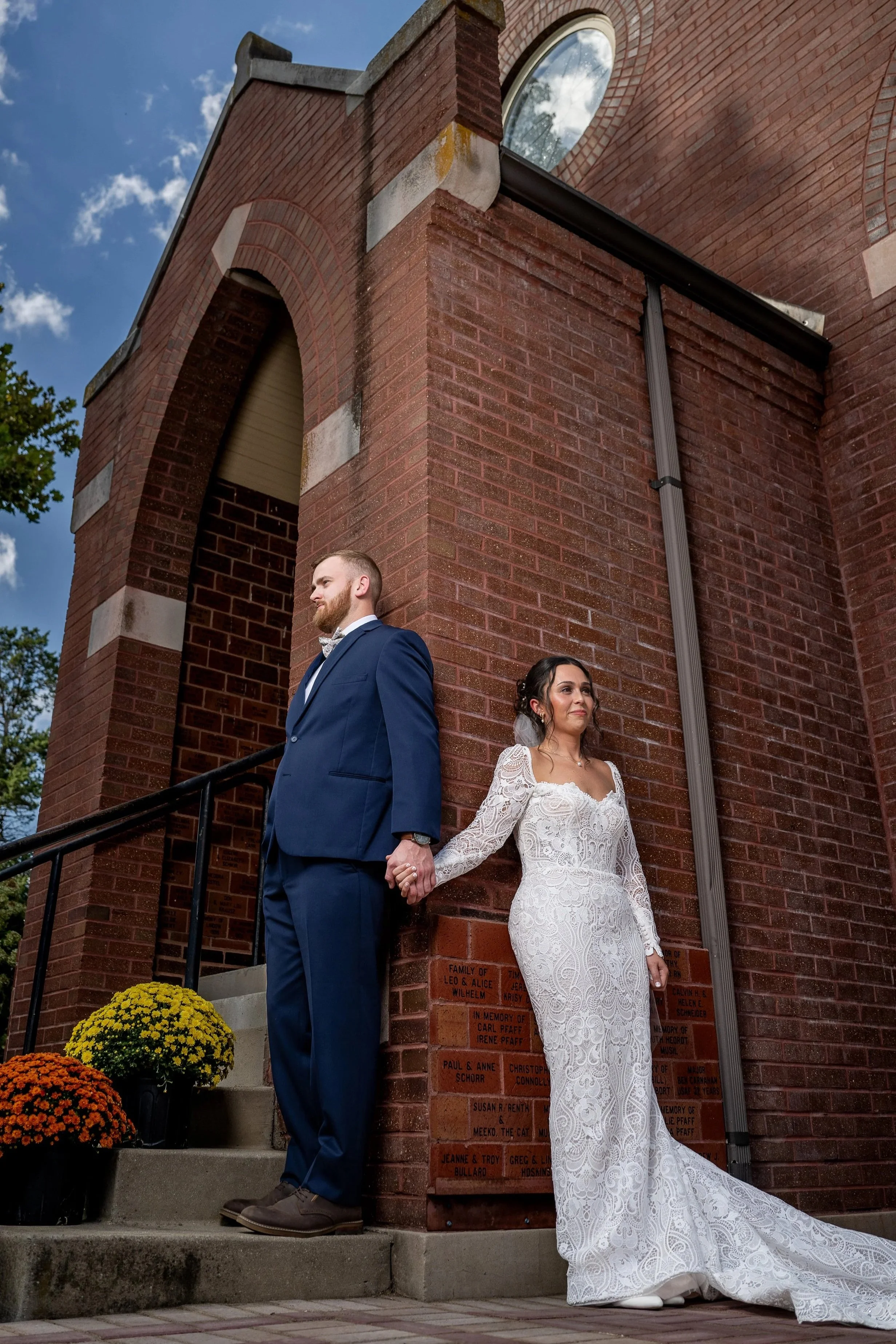 A bride and groom holding hands and standing back to back outside a brick building with a plaque, colored flowers, and cloudy sky.