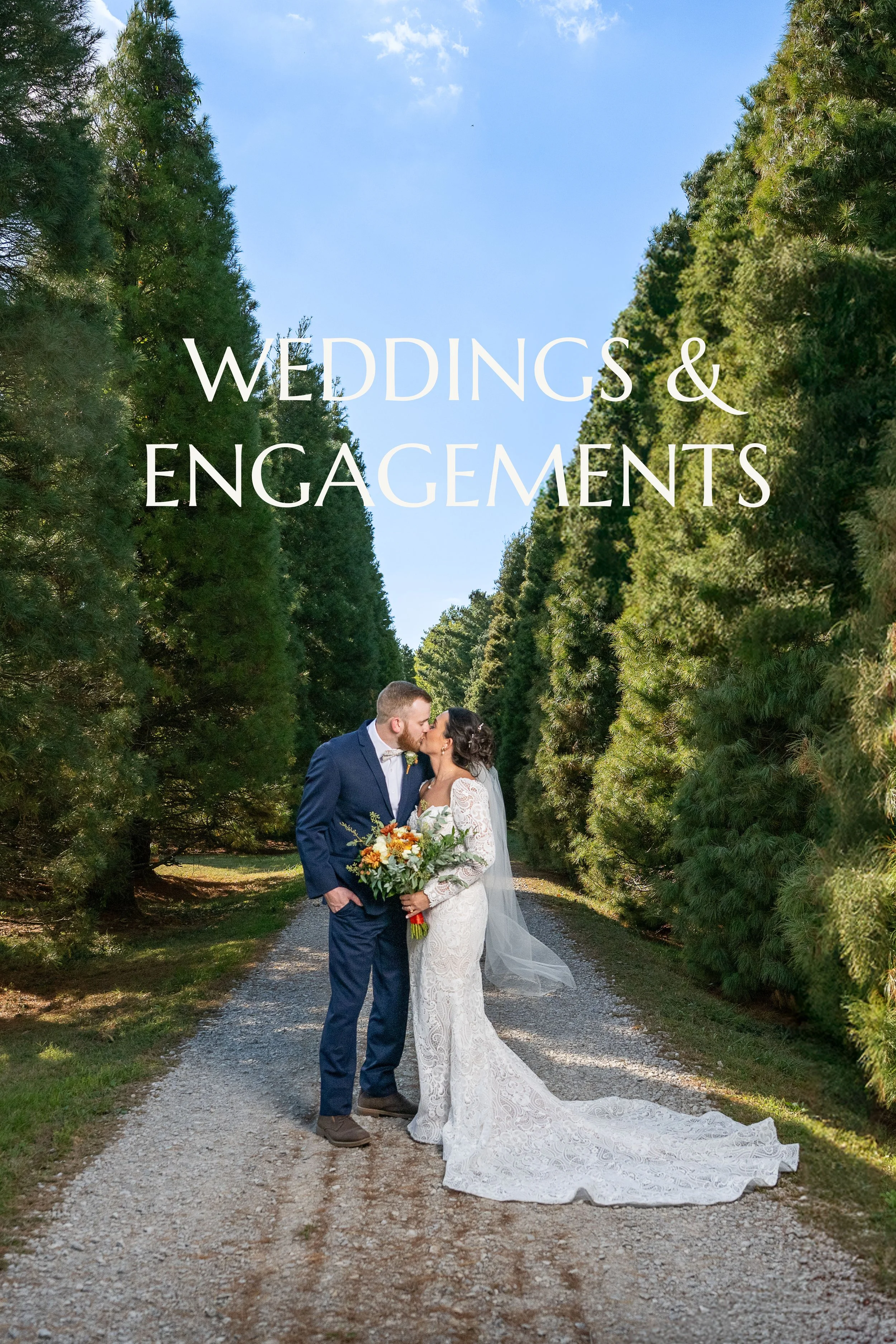 A bride and groom sharing a kiss on a gravel path surrounded by tall green trees on a sunny day.