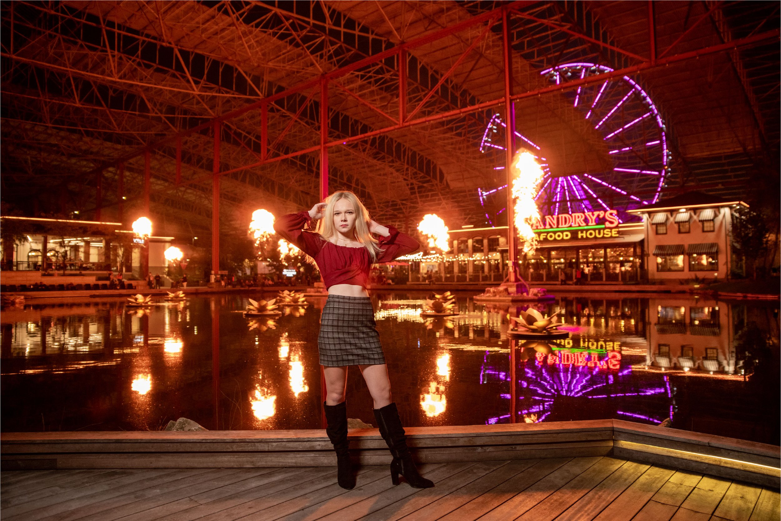 A young woman with blonde hair posing at an amusement park at night. Behind her, there are bright lights, a Ferris wheel, flames, and a sign that says 'Landry's Seafood House.' The water reflects the colorful lights and fireworks in the background.