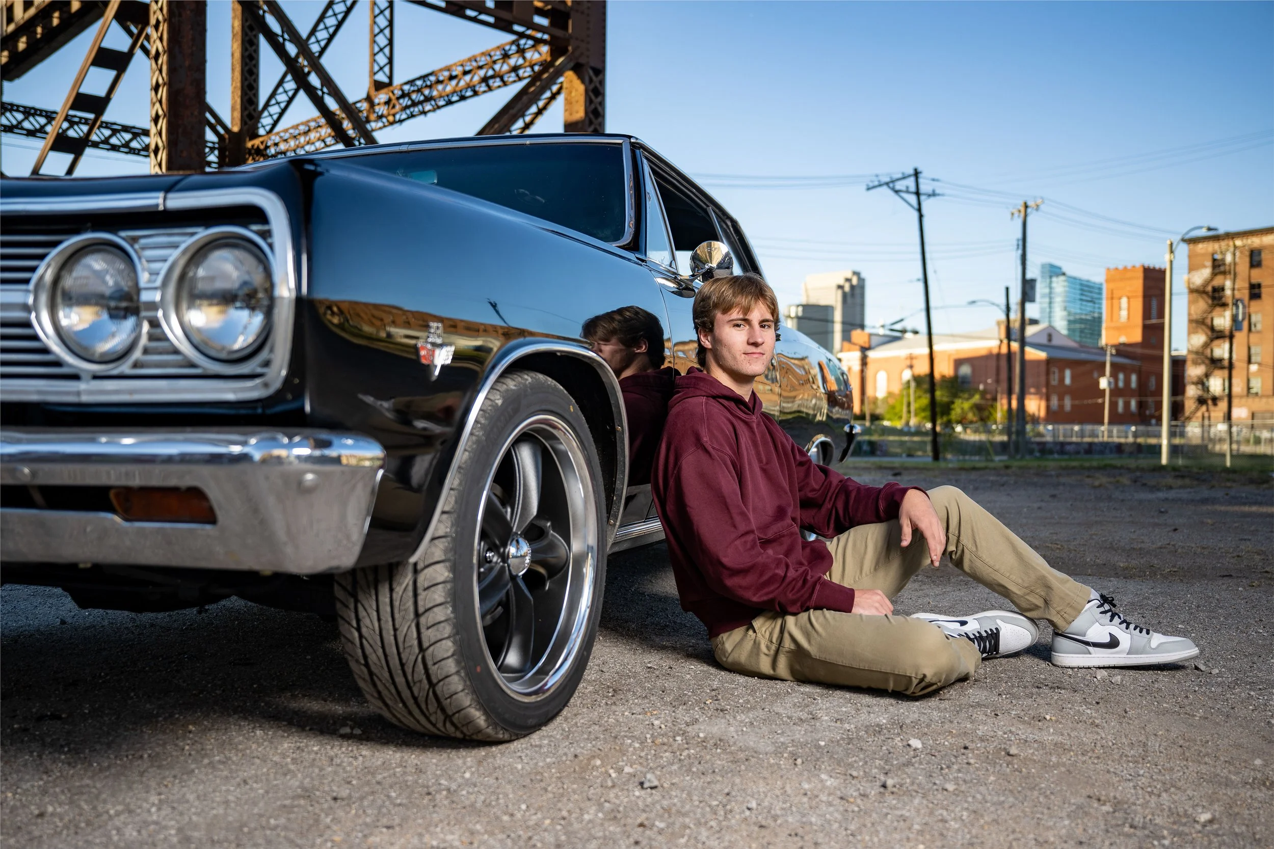 A young man with light brown hair, wearing a maroon hoodie, beige pants, and white sneakers, sitting on the ground with his back against a black vintage car in an urban parking lot during daylight. The car has a shiny black exterior and large wheels.