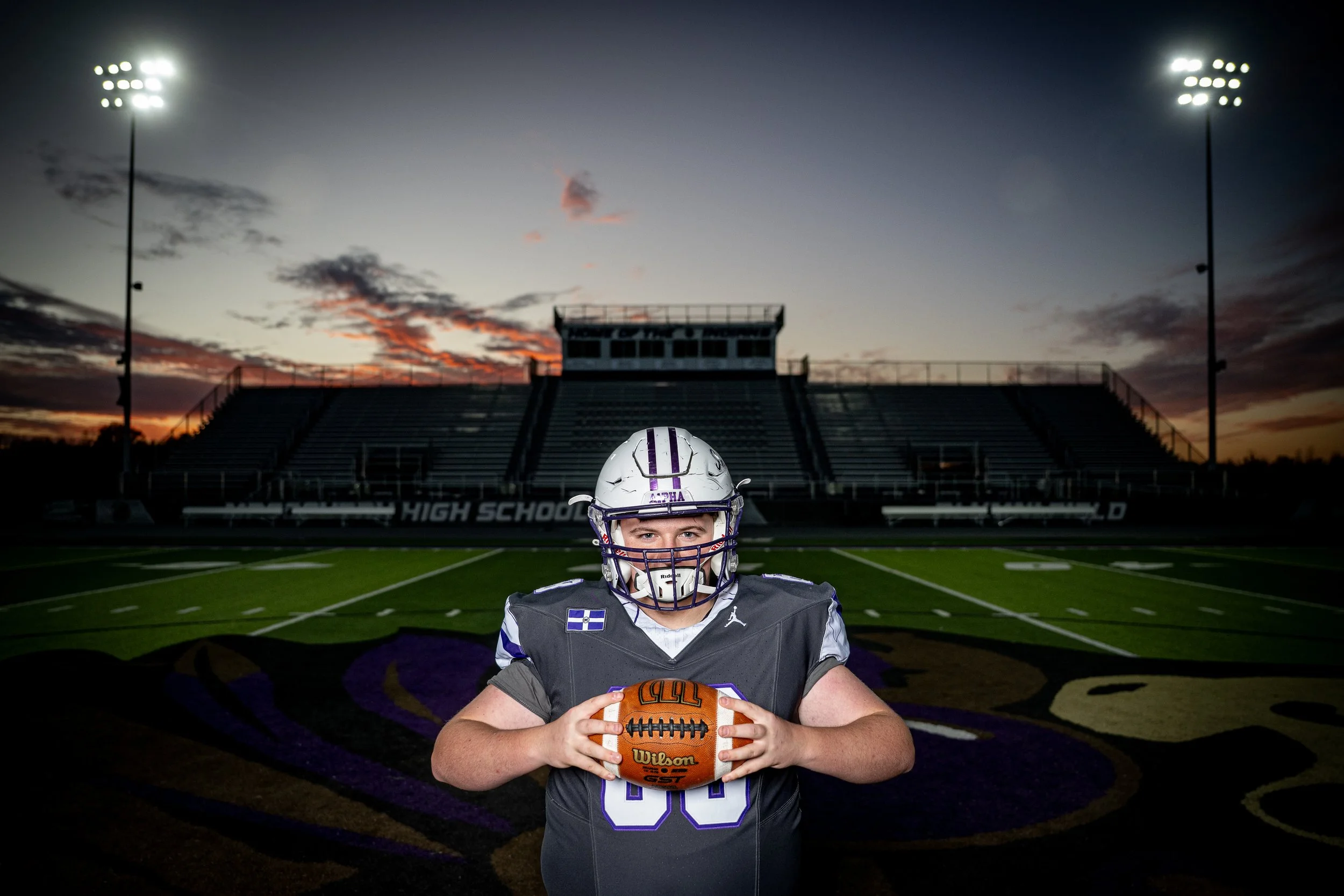 A young football player in a uniform holding a football on a football field at dusk, with stadium lights and bleachers in the background.