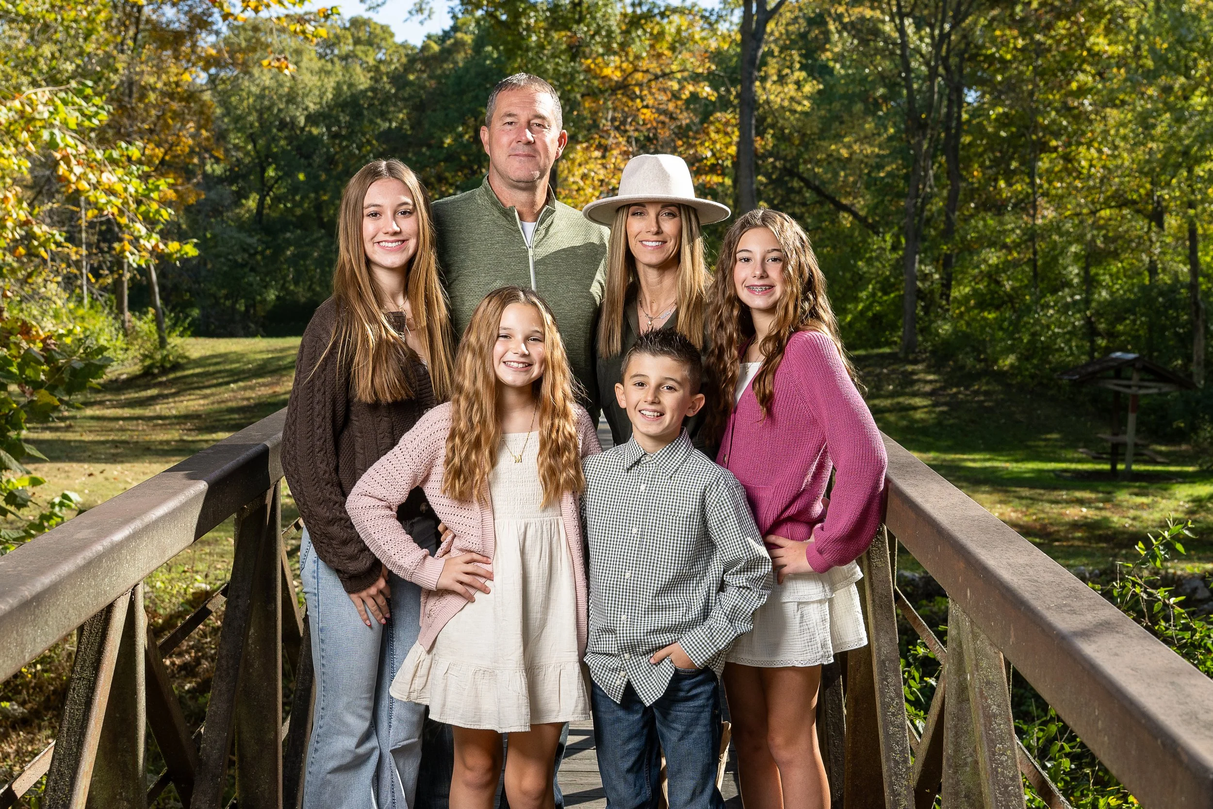 Family of seven standing on a wooden bridge in a park with green and orange fall foliage, smiling at the camera.