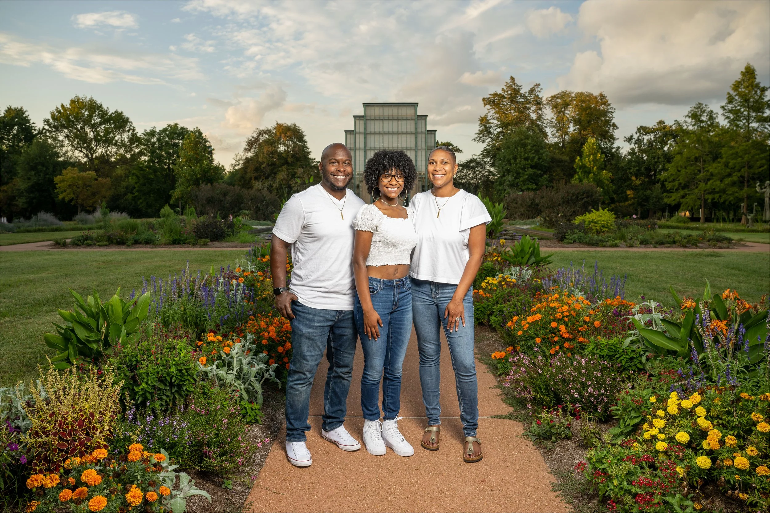 Three people standing on a garden path with flowerbeds, trees, and a glass building in the background, during daytime.