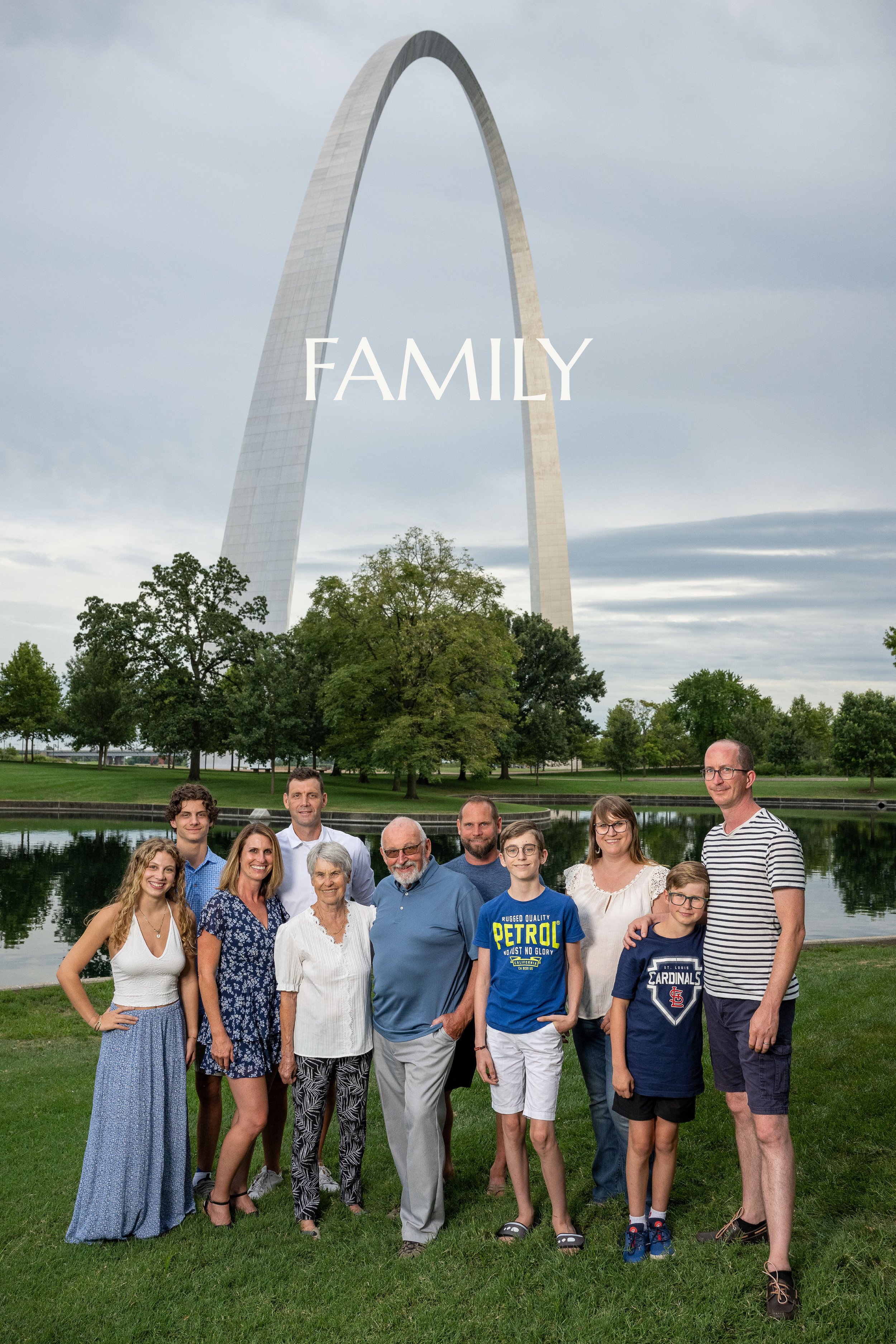 A large group of diverse people, including children, adults, and seniors, standing on a grassy area near a water with the Gateway Arch in the background on a cloudy day, with the word 'FAMILY' overlaid on the image.