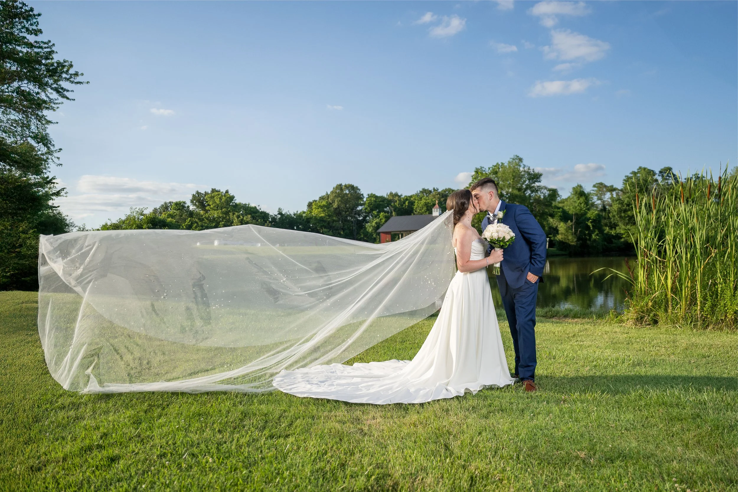 A bride and groom kiss outdoors near a lake, with the bride holding a bouquet, the bride's long veil flowing, and trees and a small building in the background.