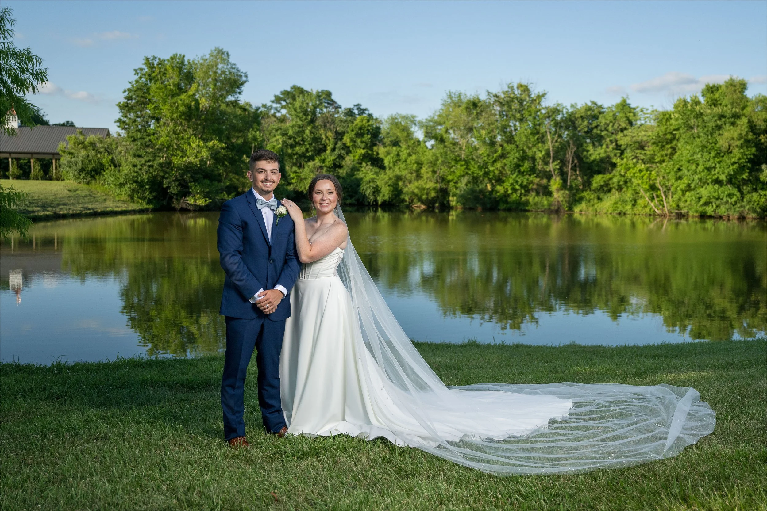 A newlywed couple standing outdoors by a lake, with green trees and blue sky in the background, smiling at the camera.