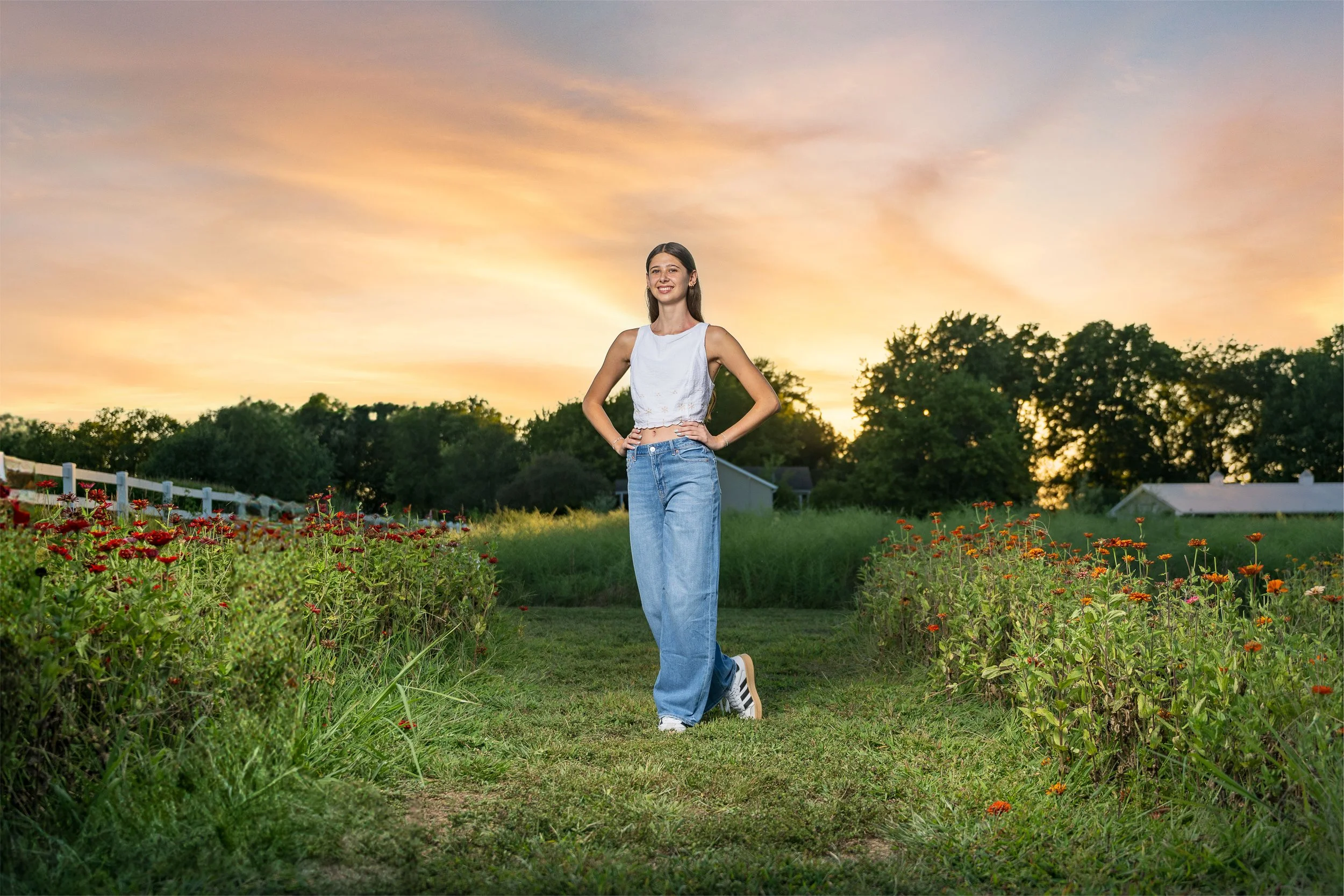A young woman standing in a flower garden during sunset with trees and small buildings in the background.