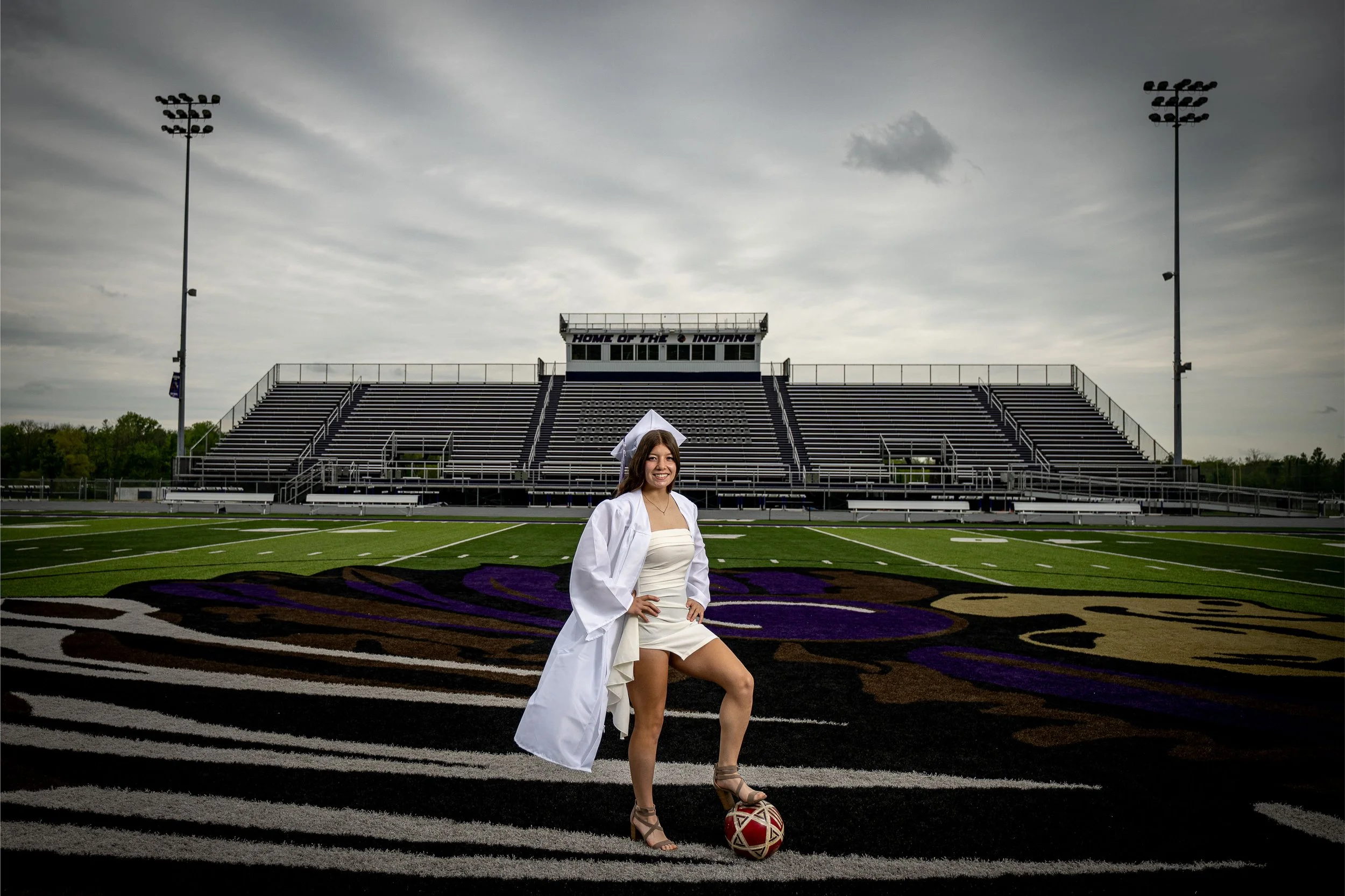 A young woman in a white graduation gown standing on a football field with a soccer ball, with stadium bleachers and floodlights in the background.