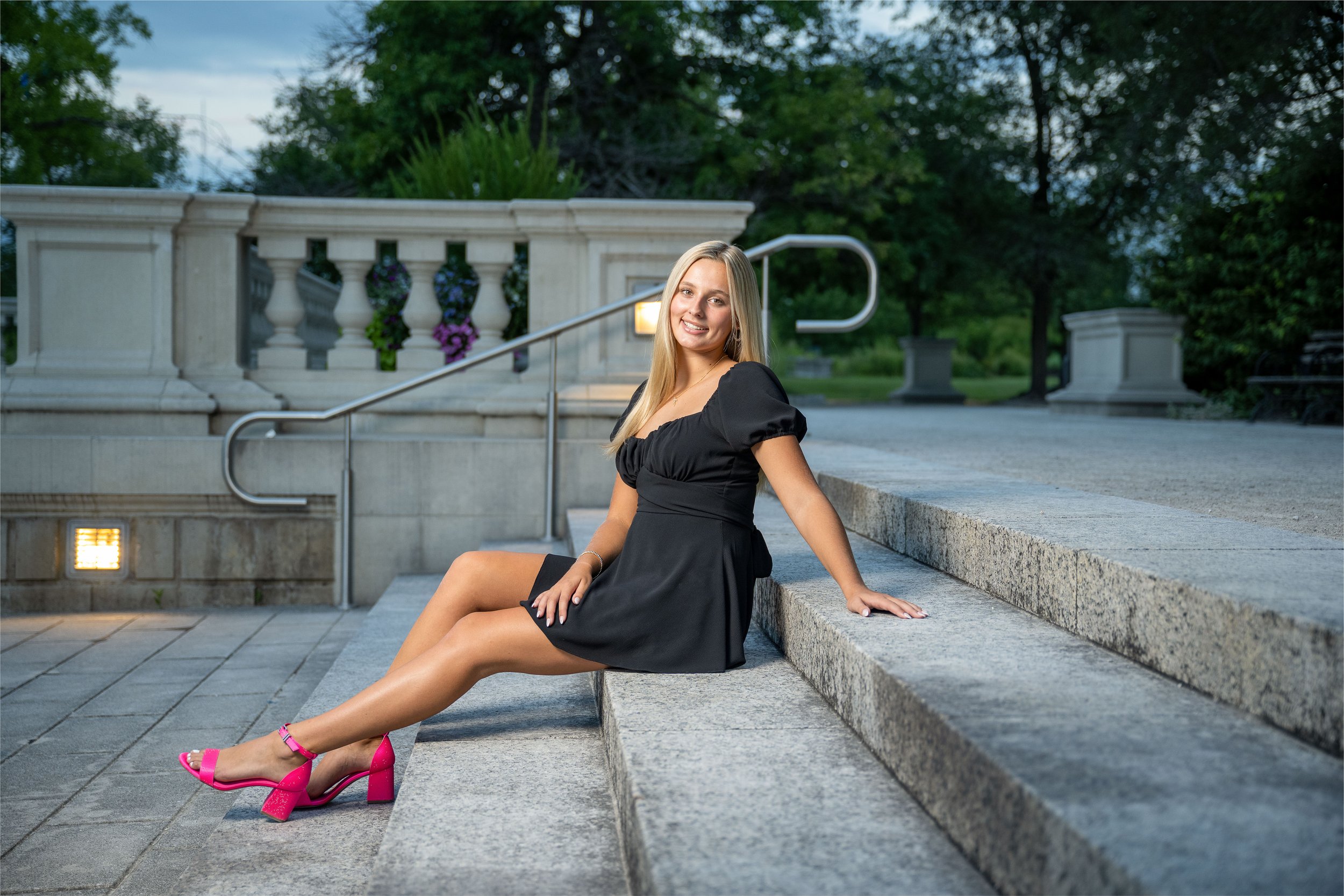 A young woman with long blonde hair sitting on stone steps outdoors, smiling at the camera, wearing a black dress and bright pink high-heeled shoes, with greenery and decorative planters in the background.